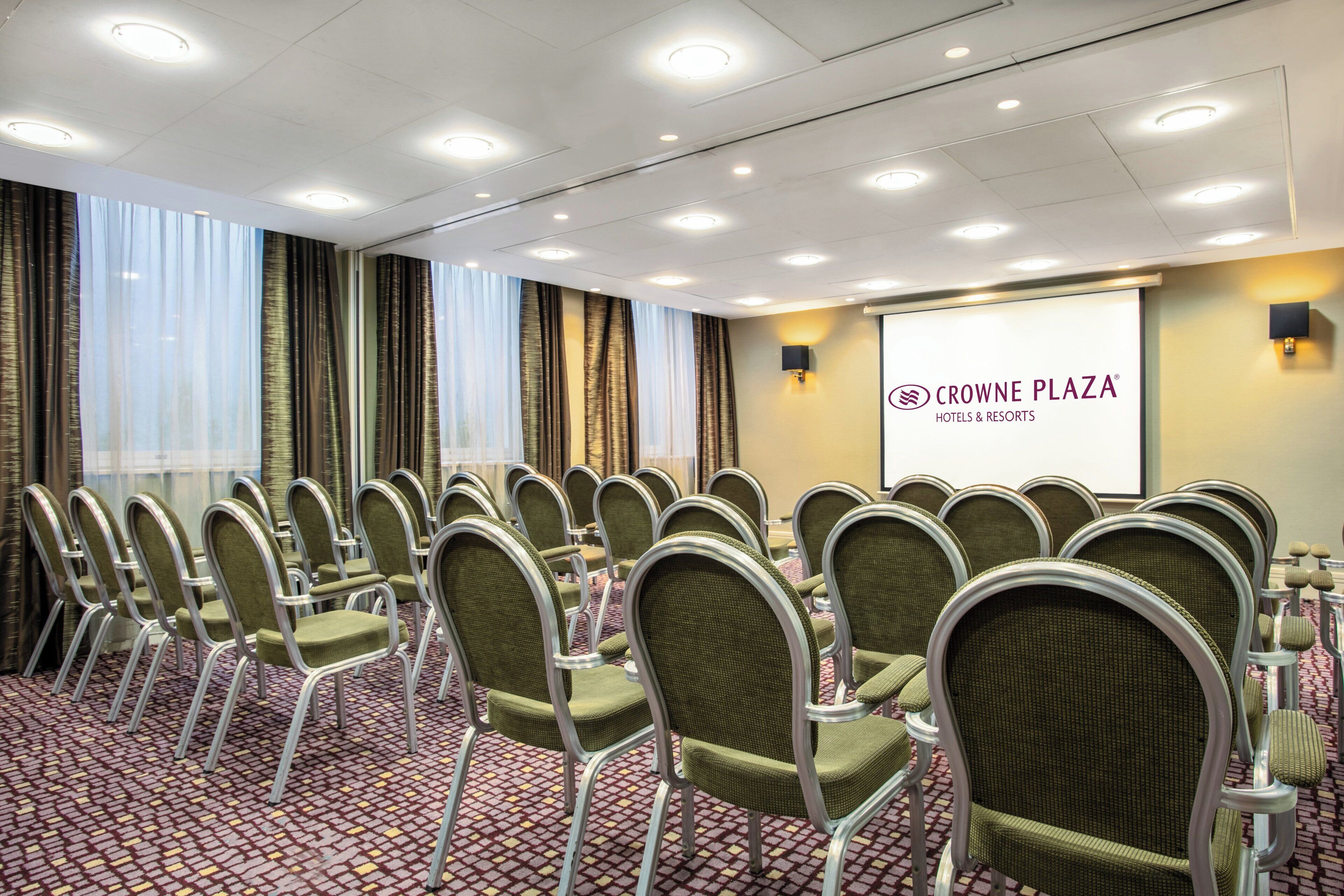 A conference room with rows of chairs, a projector screen, and a logo for Crowne Plaza Hotels & Resorts.