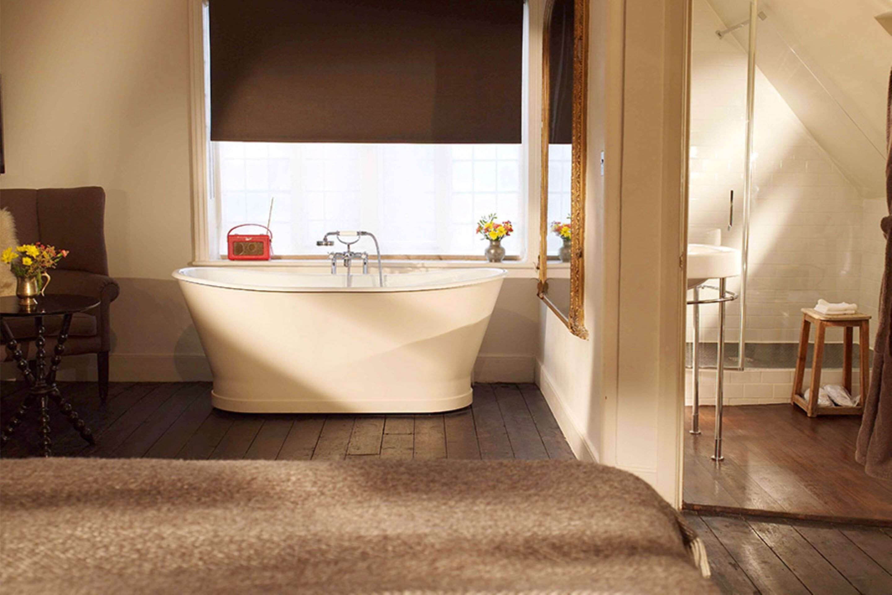 Une salle de bain avec une baignoire sur pieds, une fenêtre avec un store, une petite table avec un vase et un miroir au mur.