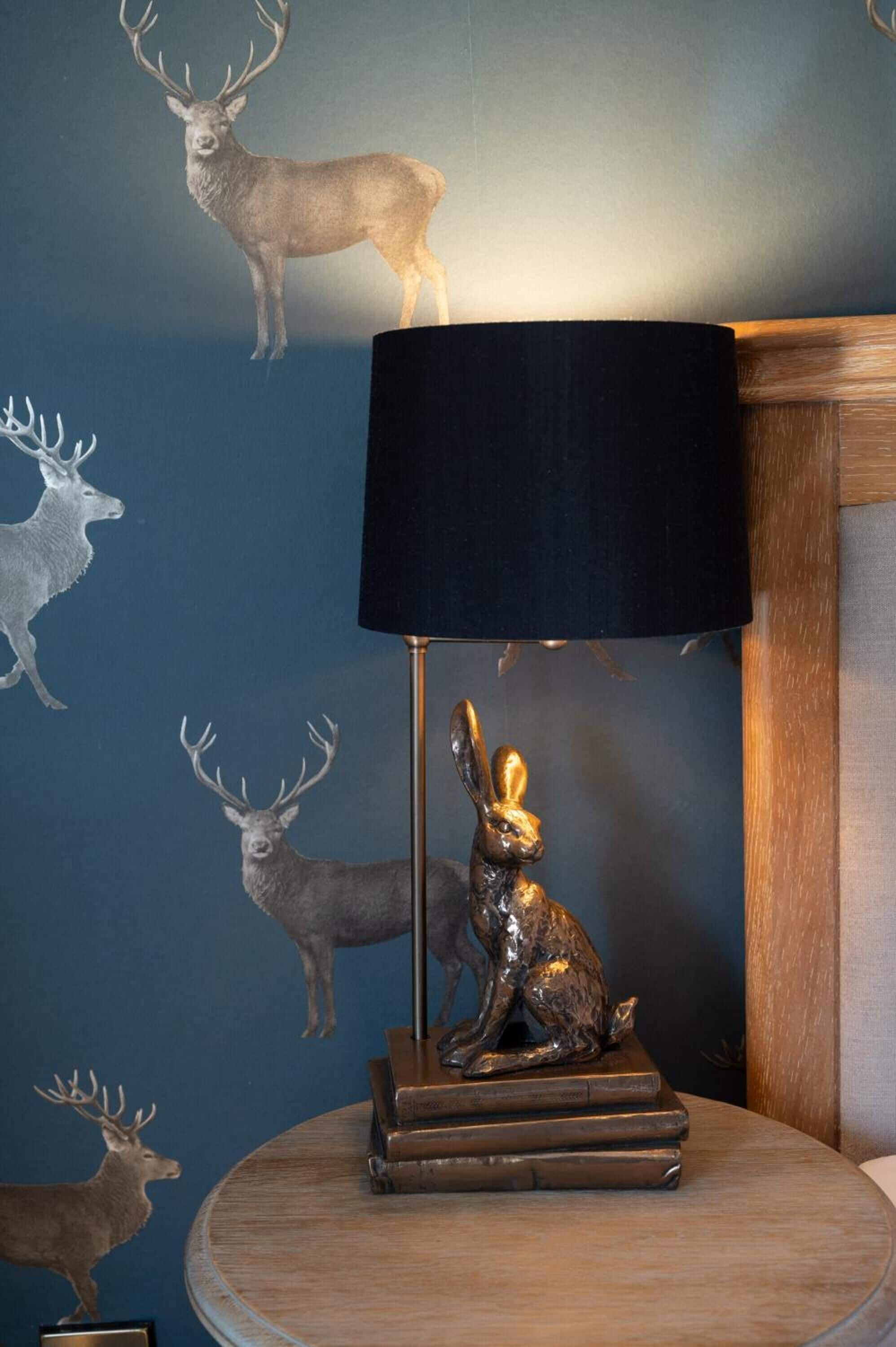 A lamp with a black shade on a wooden table, surrounded by deer and rabbit wall decals.