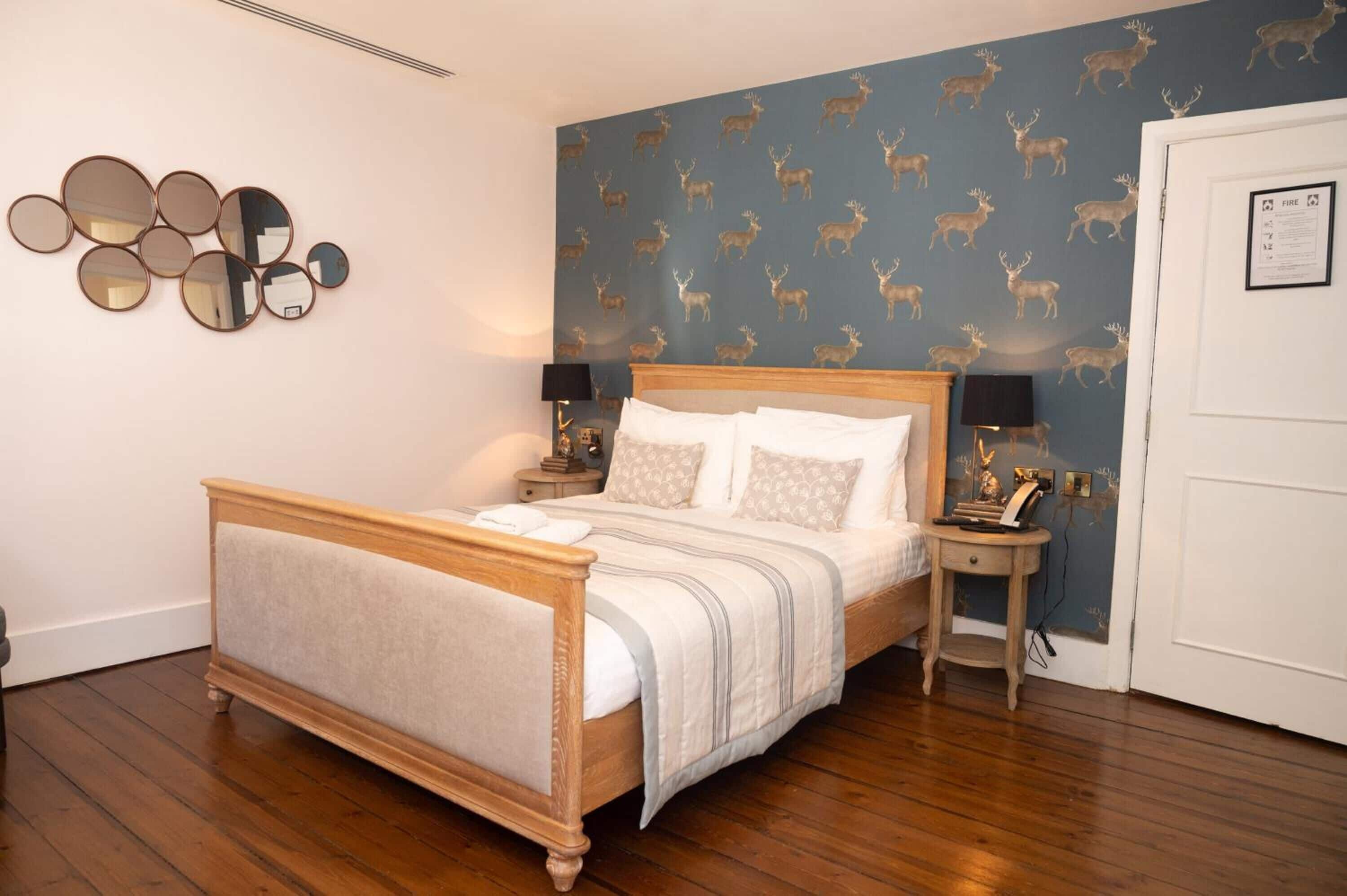 A neatly made bed with a wooden frame and white bedding, a wall with a deer pattern wallpaper, and a side table with a lamp and a framed picture.