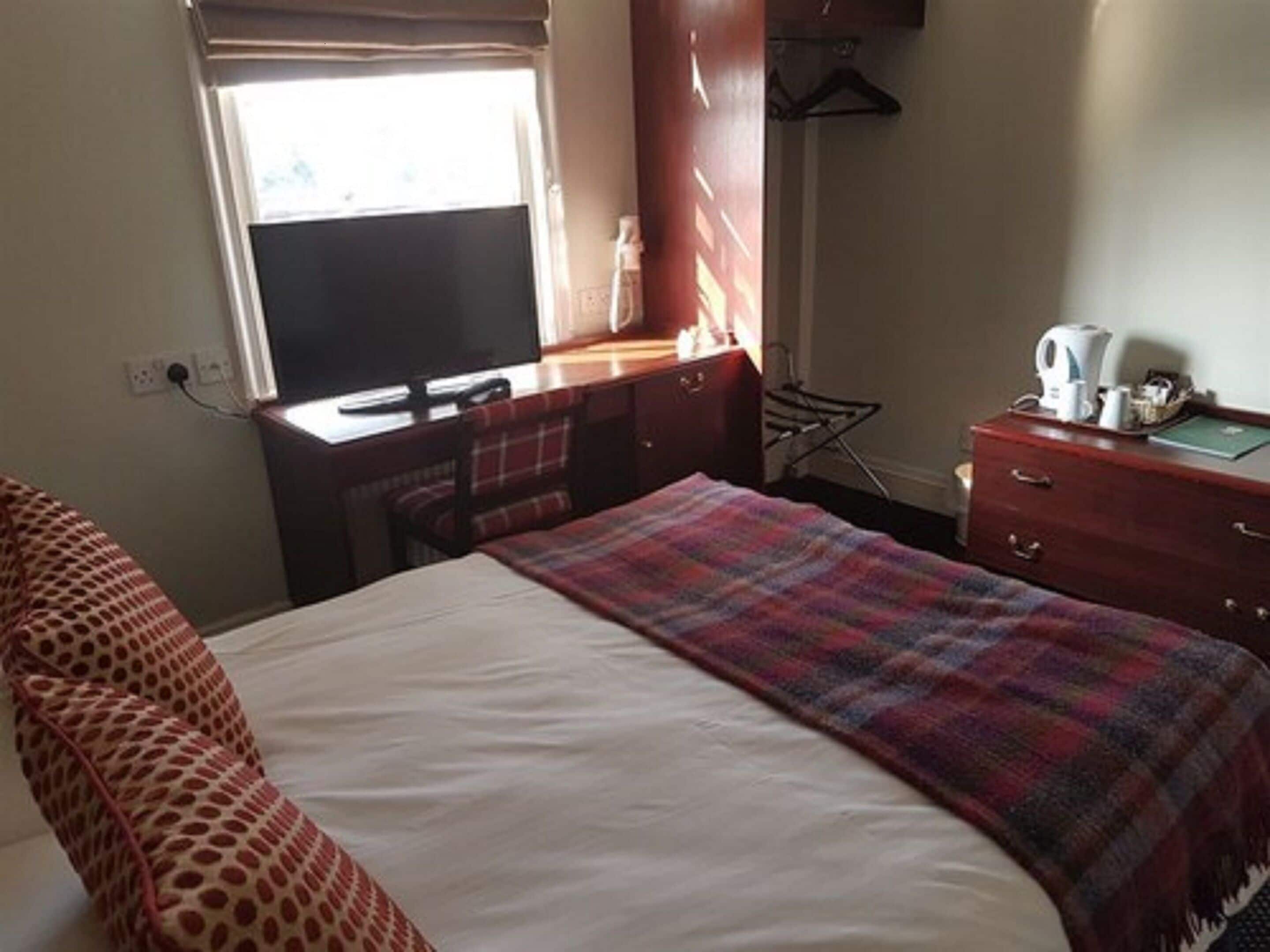 Une chambre d’hôtel comprenant un lit, un bureau avec un ordinateur, une chaise, une commode et une fenêtre avec des rideaux.