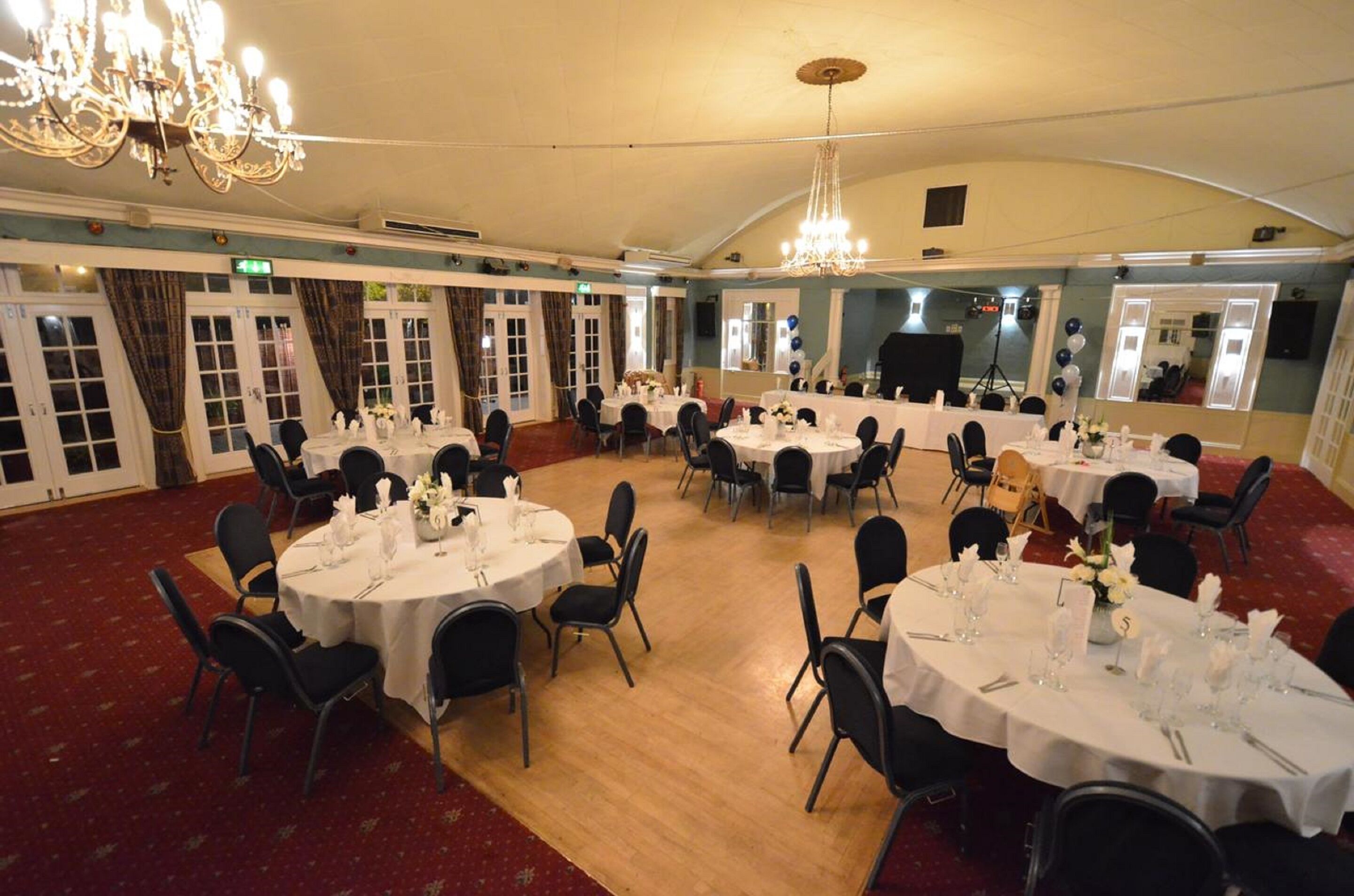 Un salón de banquetes con mesas redondas dispuestas para cenar, sillas negras, candelabros y un suelo cubierto de alfombra roja.
