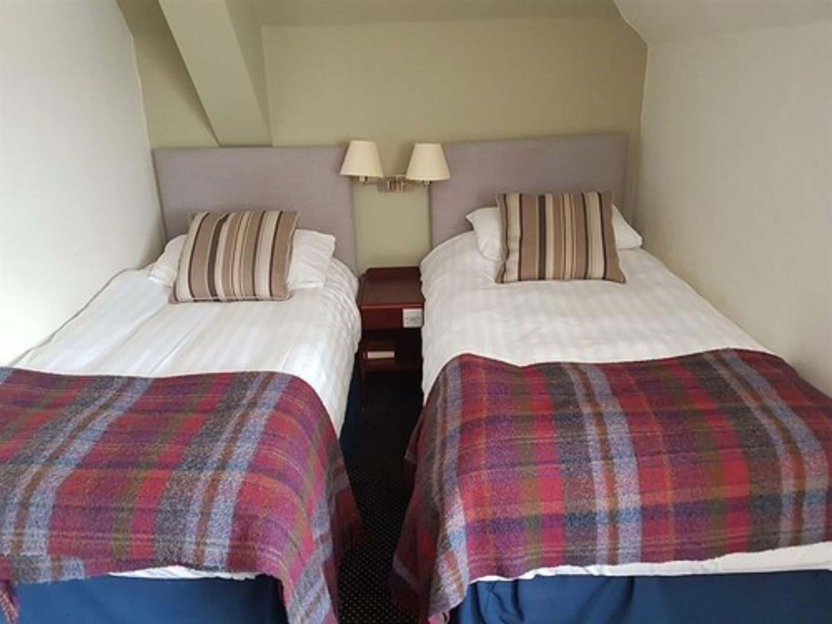 A room with two single beds, each with a plaid blanket and pillows. There are two bedside lamps and a nightstand between the beds.