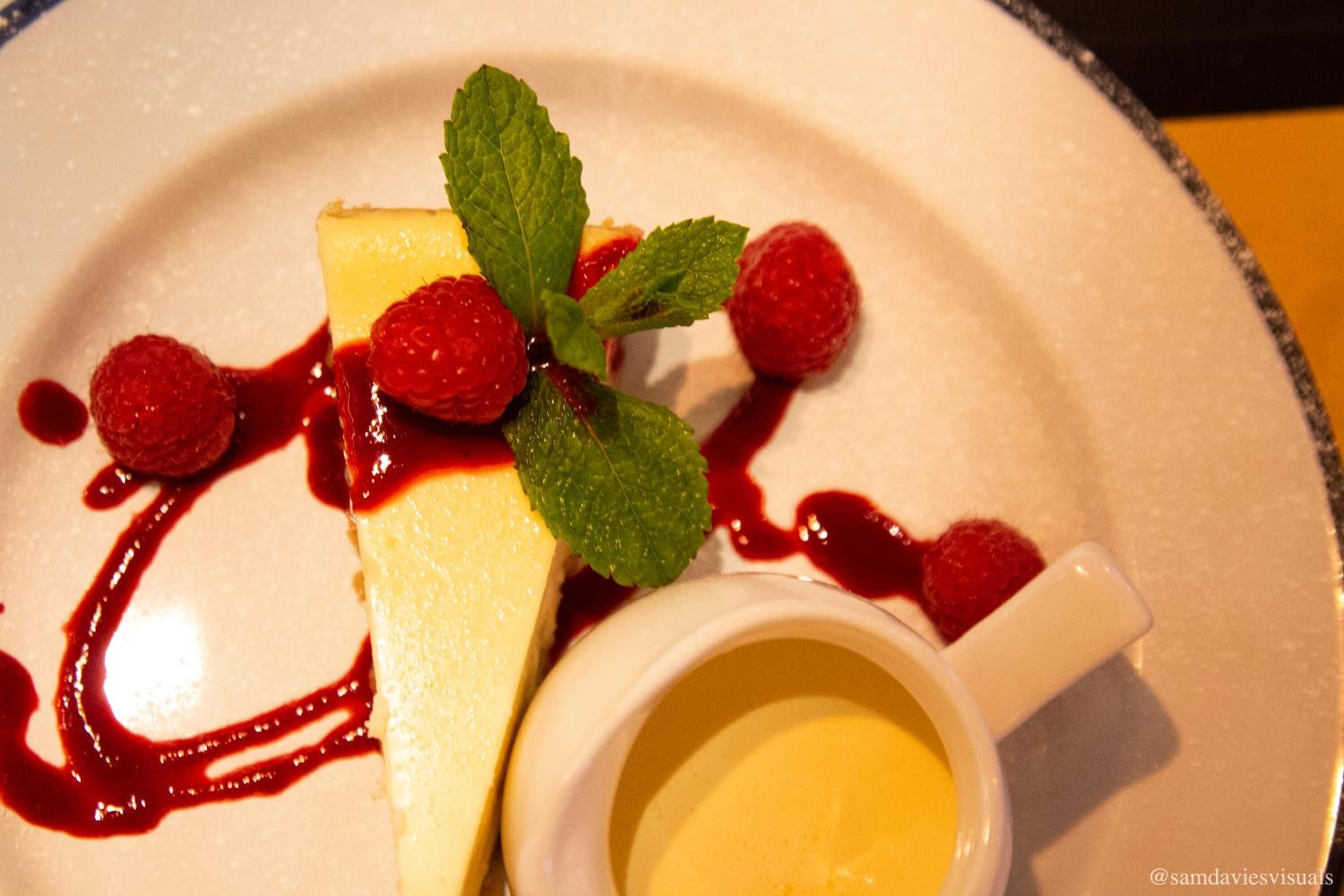 Un plato de postre con pastel de queso, frambuesas, menta y salsa.