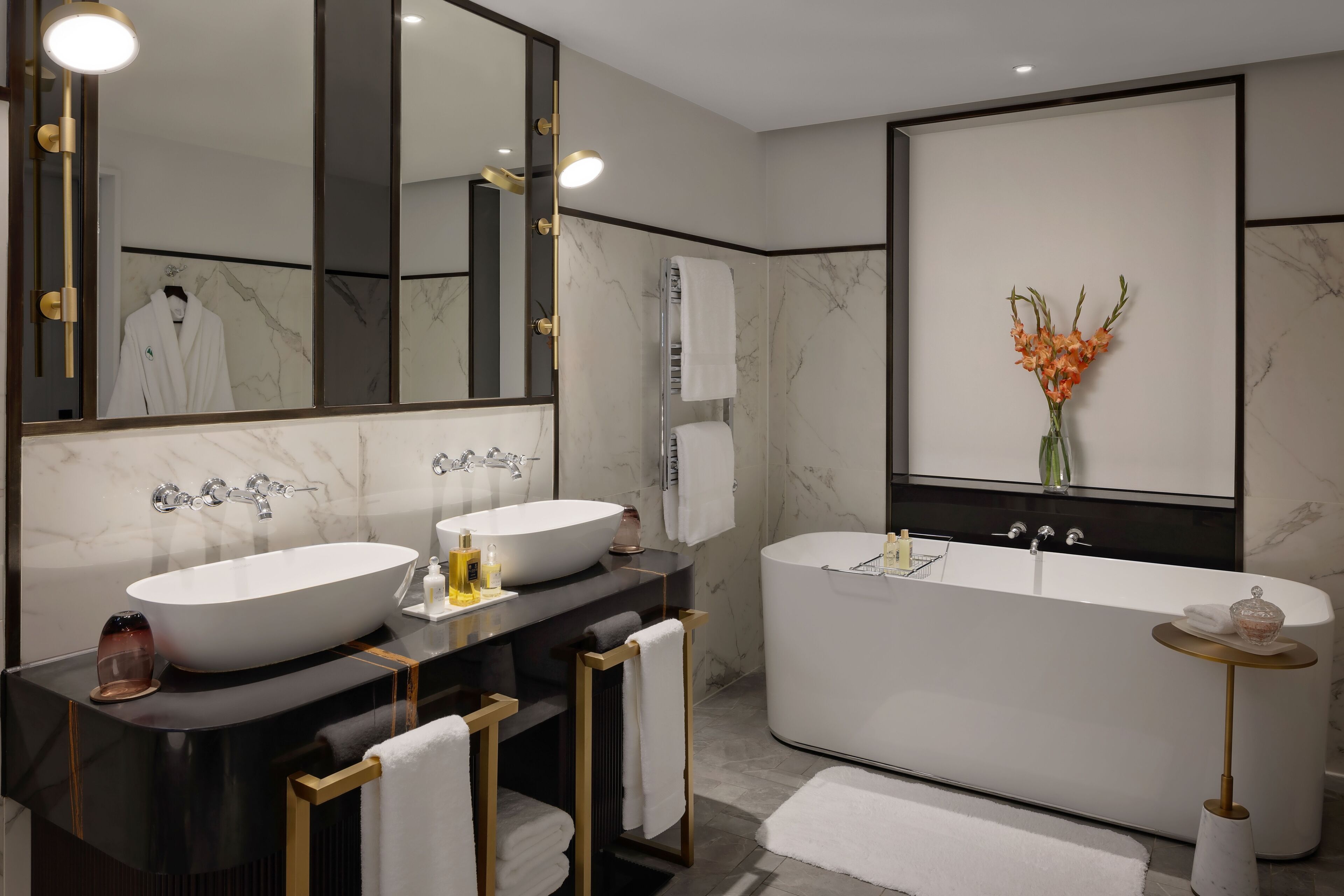 A modern bathroom with a large oval sink, a freestanding bathtub, and a large mirror. There are two towel racks and a small round table with a vase of flowers.