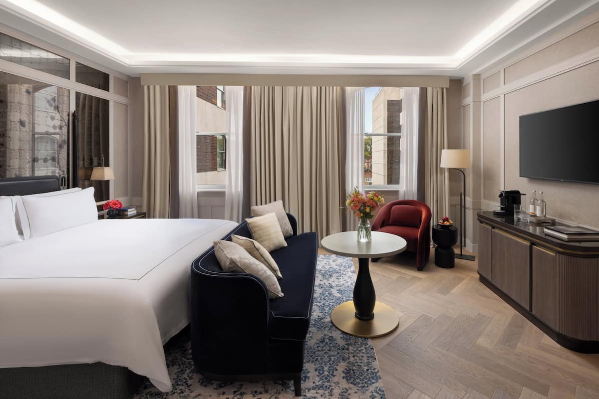 A hotel room with a large bed, a sofa, a small table with flowers, a TV, and a minibar.