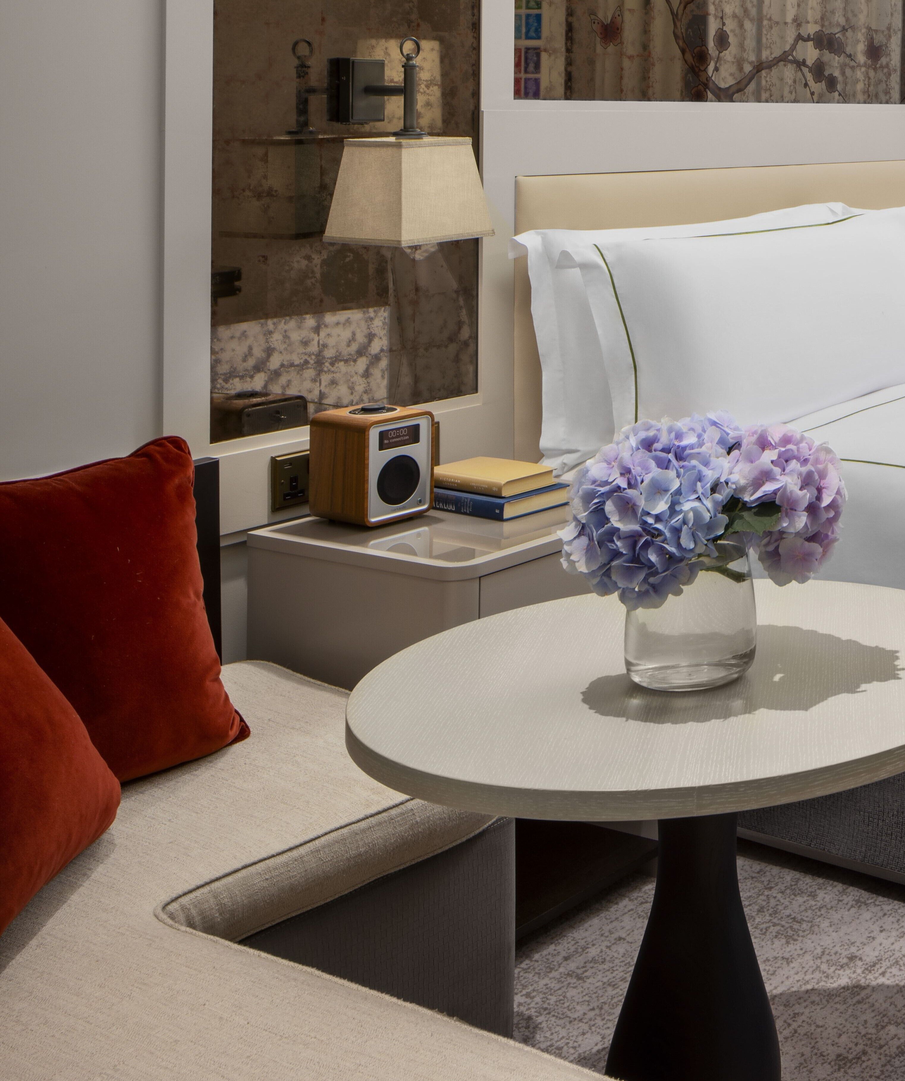 A modern hotel room with a round table, a red pillow, a wooden radio, and a vase with flowers.