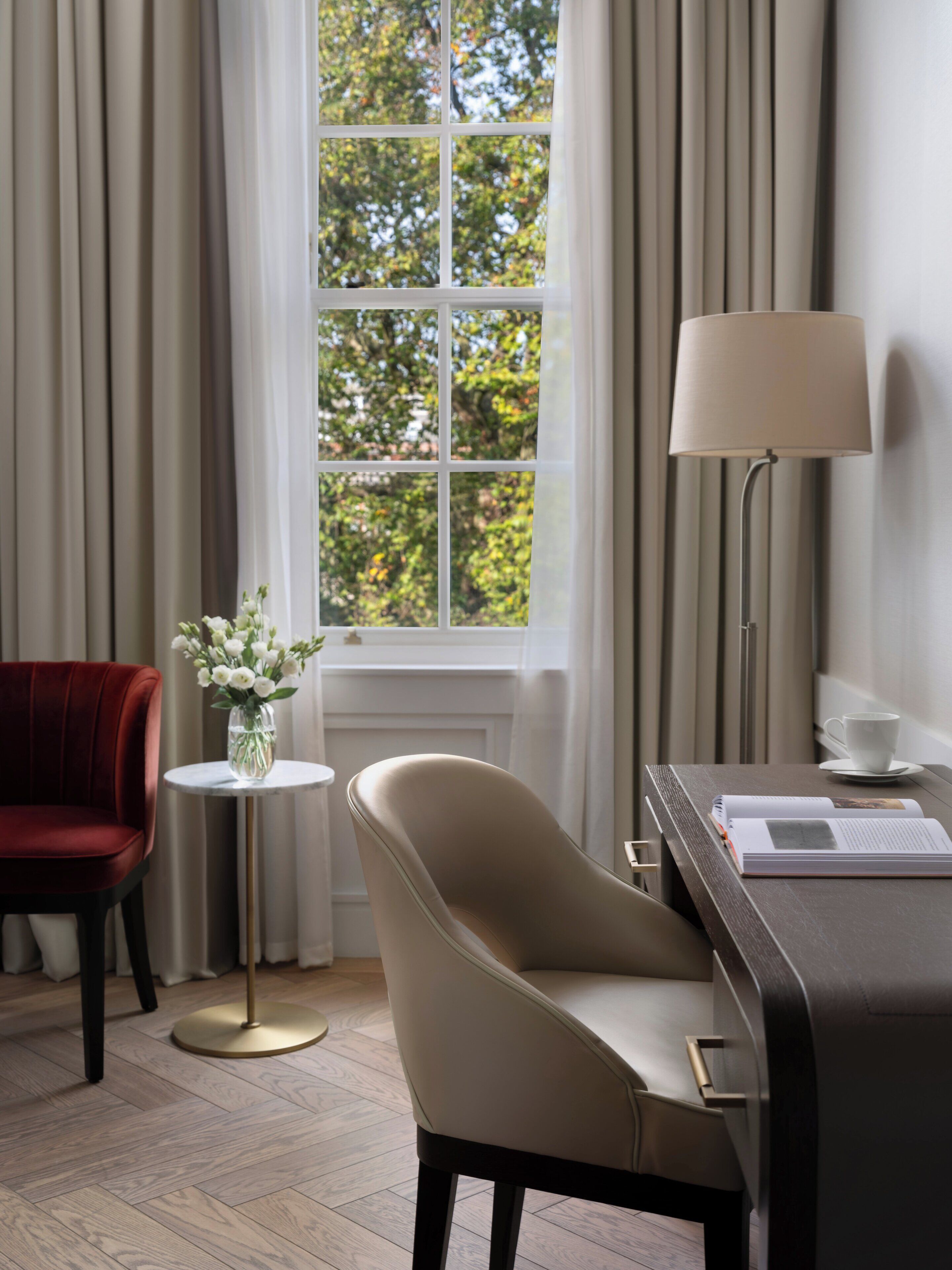 Una habitación con un ventanal grande, una silla roja, un sillón color beige, una pequeña mesa redonda con un jarrón de flores, una lámpara y un libro sobre un escritorio.
