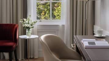 Una habitación con un ventanal grande, una silla roja, un sillón color beige, una pequeña mesa redonda con un jarrón de flores, una lámpara y un libro sobre un escritorio.