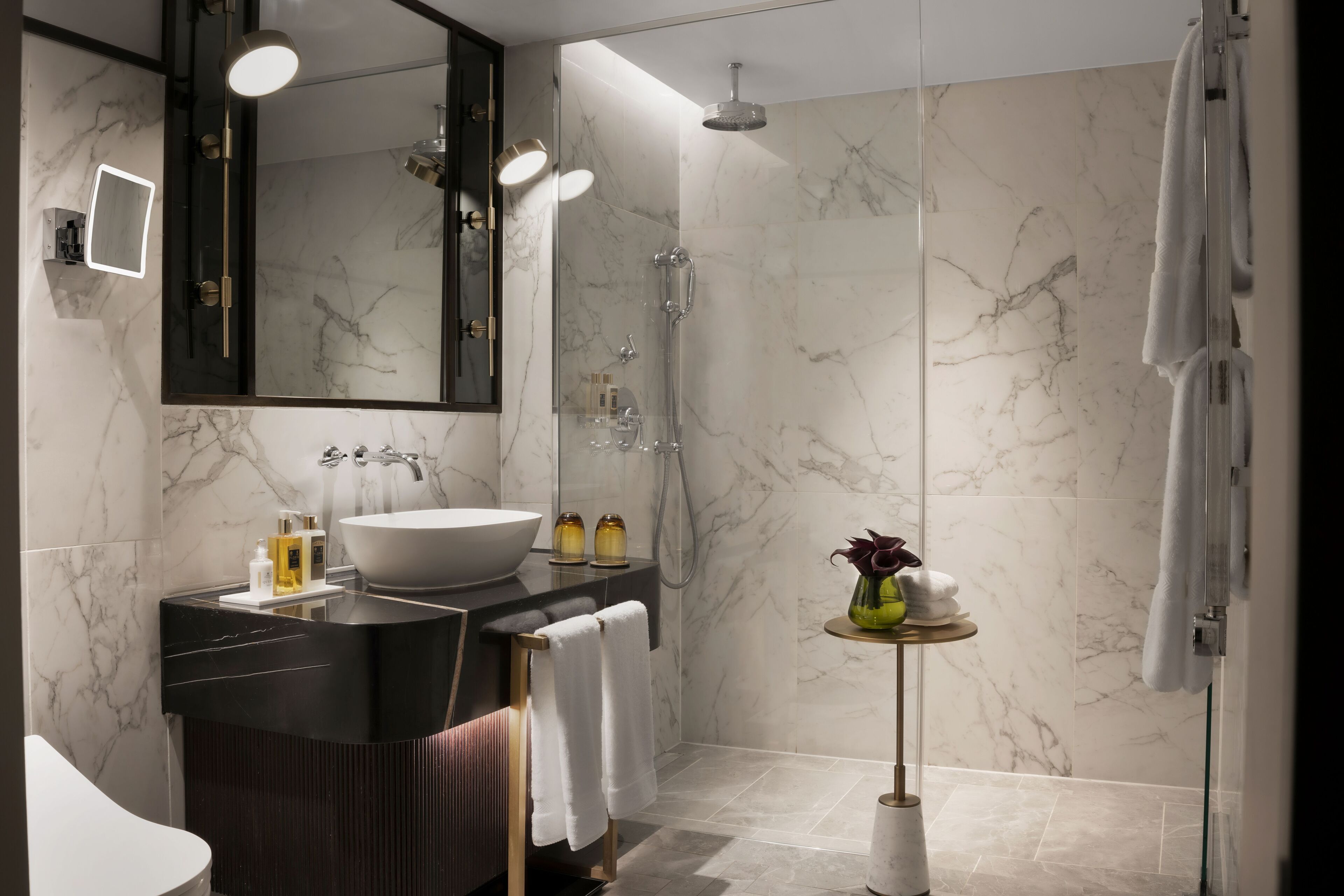Un baño moderno con un espejo grande, un lavabo sobre encimera, ducha con mampara de vidrio y una mesita redonda con artículos de aseo.