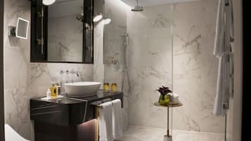 Un baño moderno con un espejo grande, un lavabo sobre encimera, ducha con mampara de vidrio y una mesita redonda con artículos de aseo.
