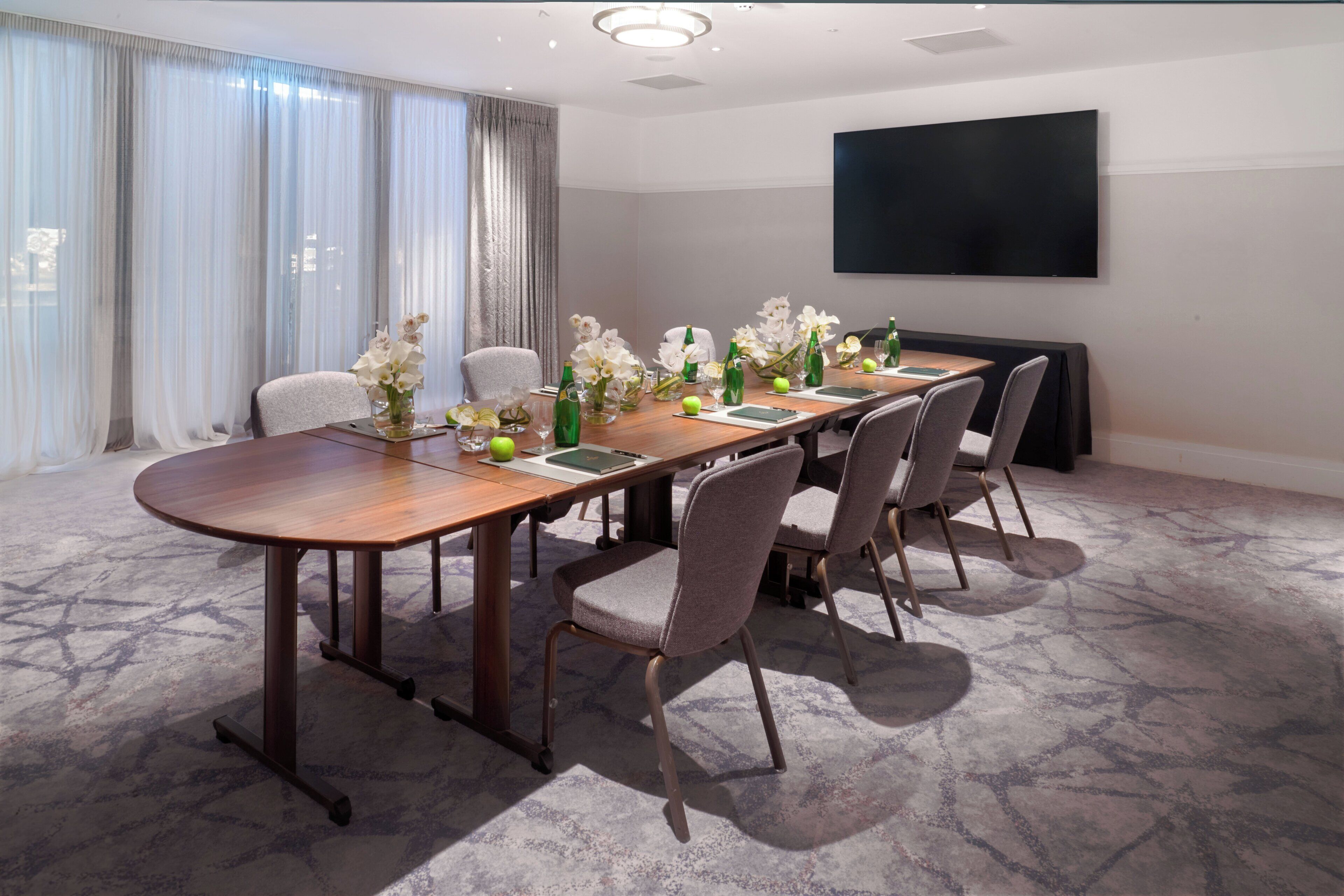 A conference room with a long table, six chairs, and a flat-screen TV on the wall.