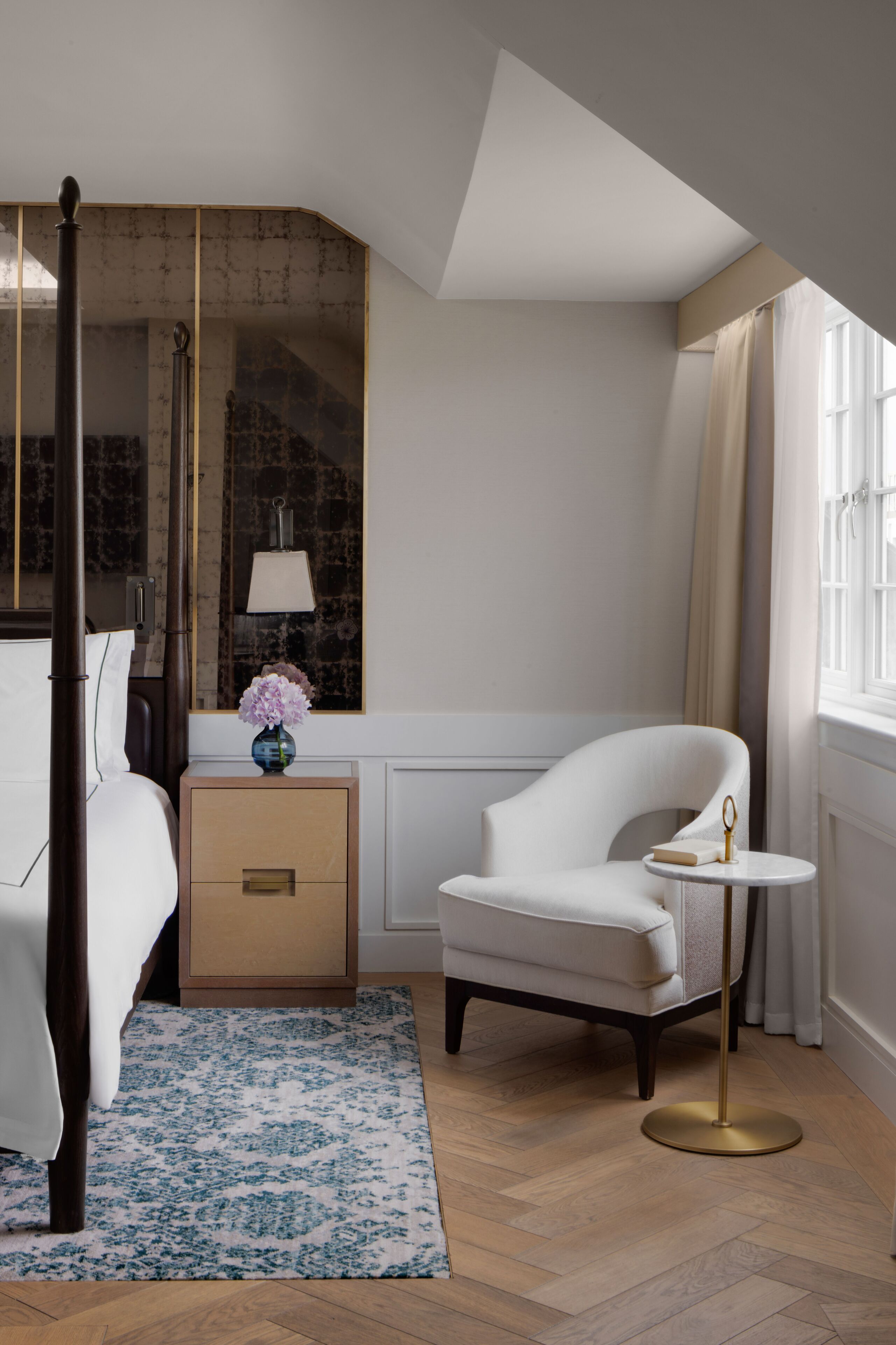 Een slaapkamer met een hemelbed, een nachtkastje met een lamp, een stoel en een kleine ronde tafel.