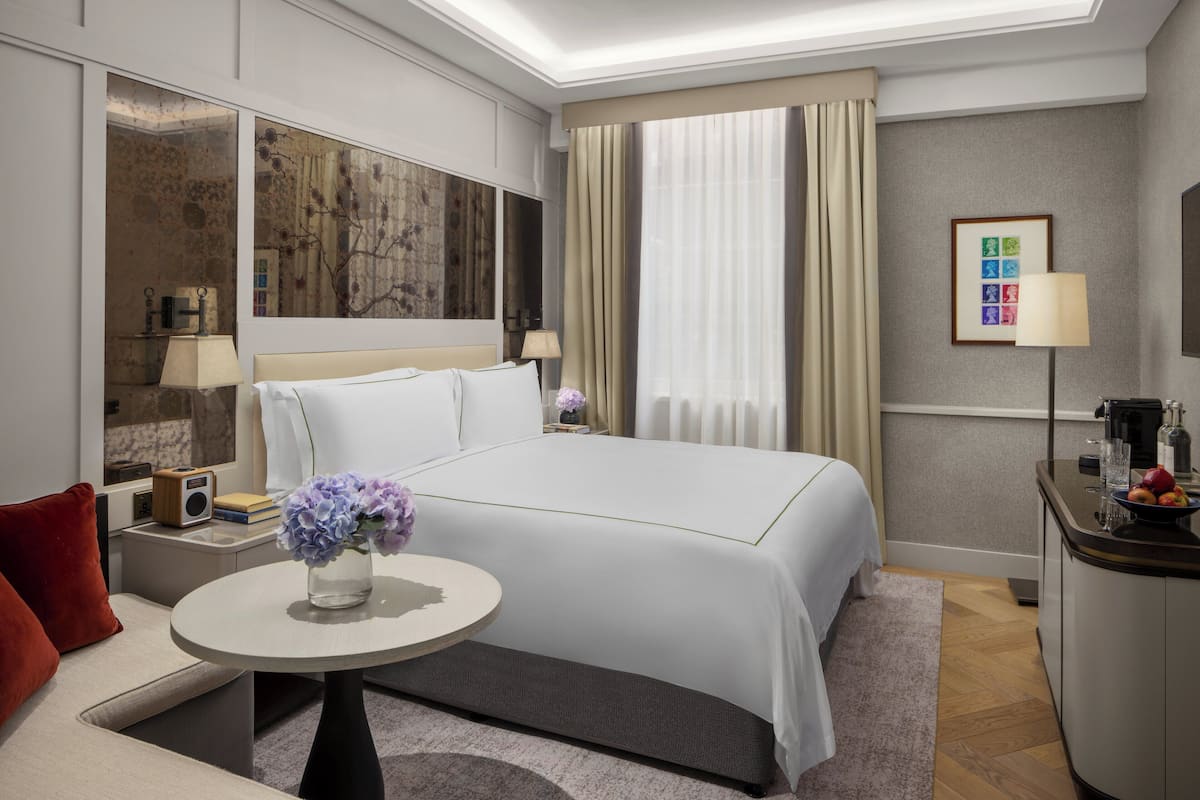 A hotel room with a large bed, a small round table with a vase of flowers, a sofa, a desk with a lamp, and a large window with curtains.