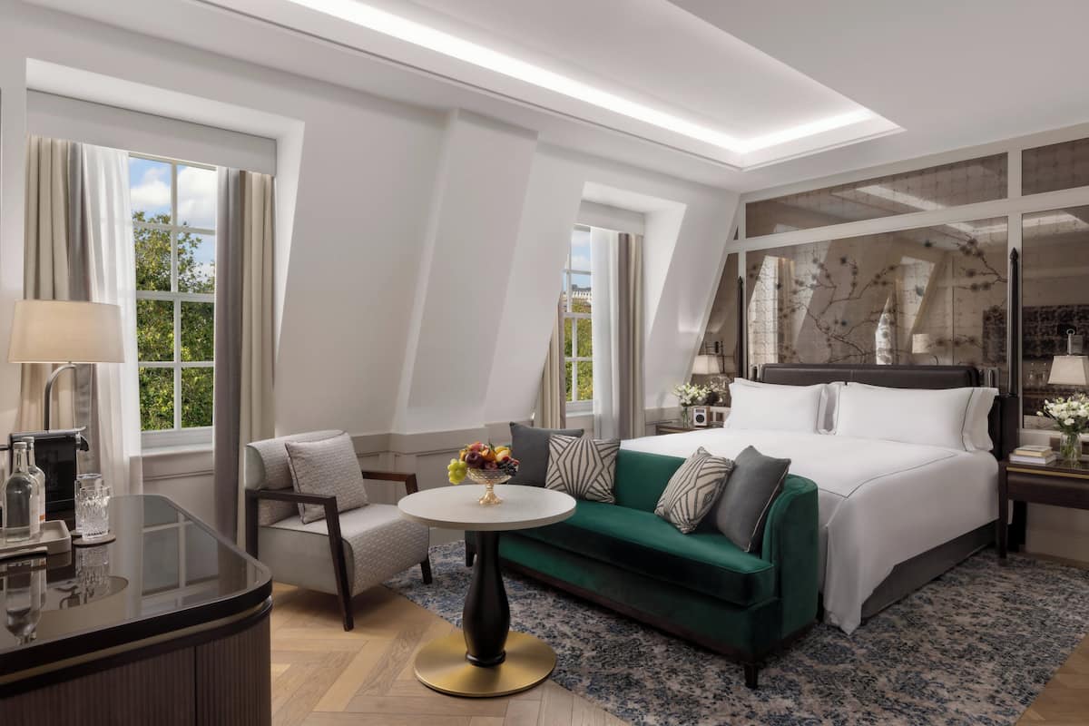 A modern hotel room with a large bed, a green sofa, a round table with a vase of flowers, and a desk with a lamp.