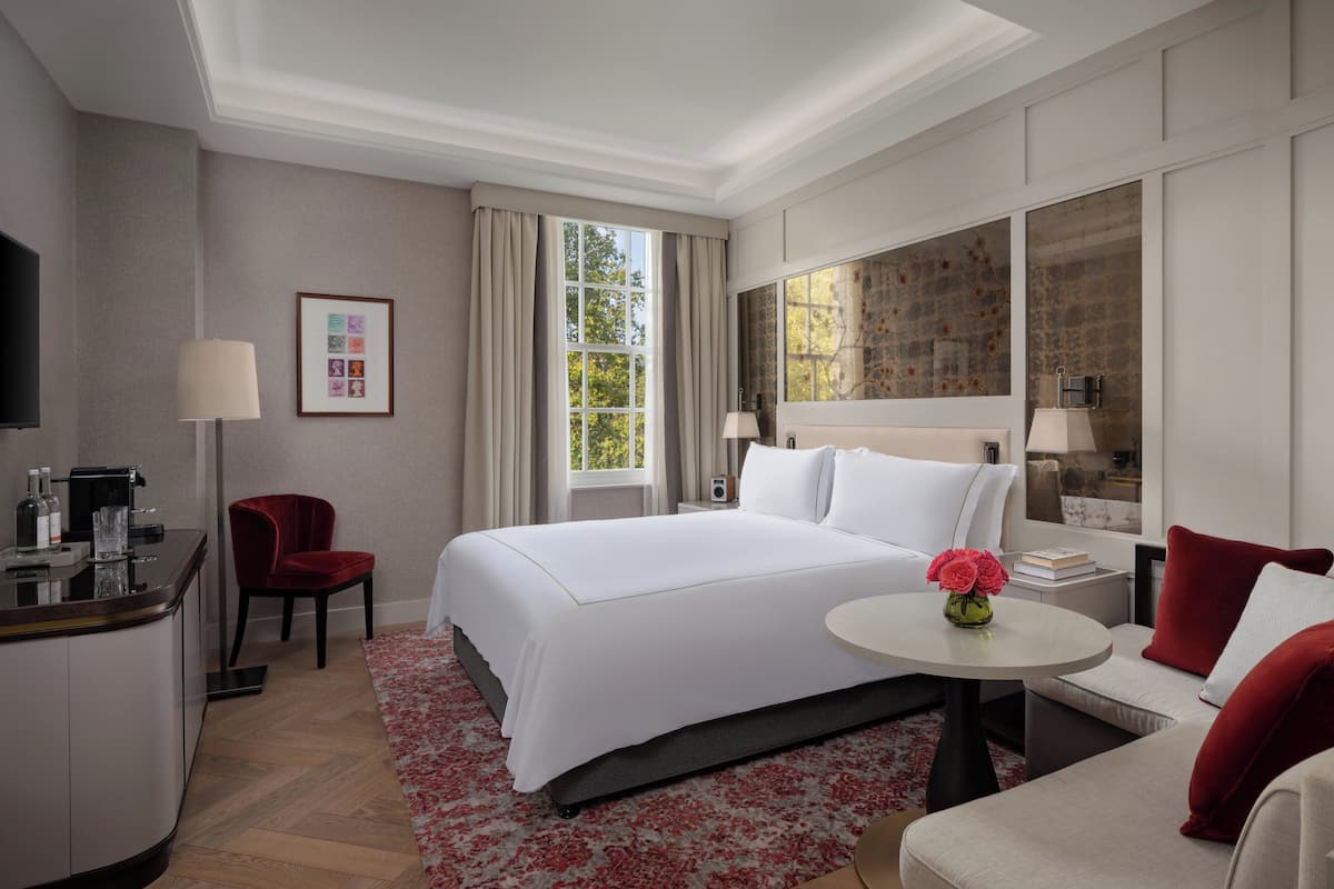 A hotel room with a large bed, a desk with a lamp, a chair, a small table with a vase, and a large window.
