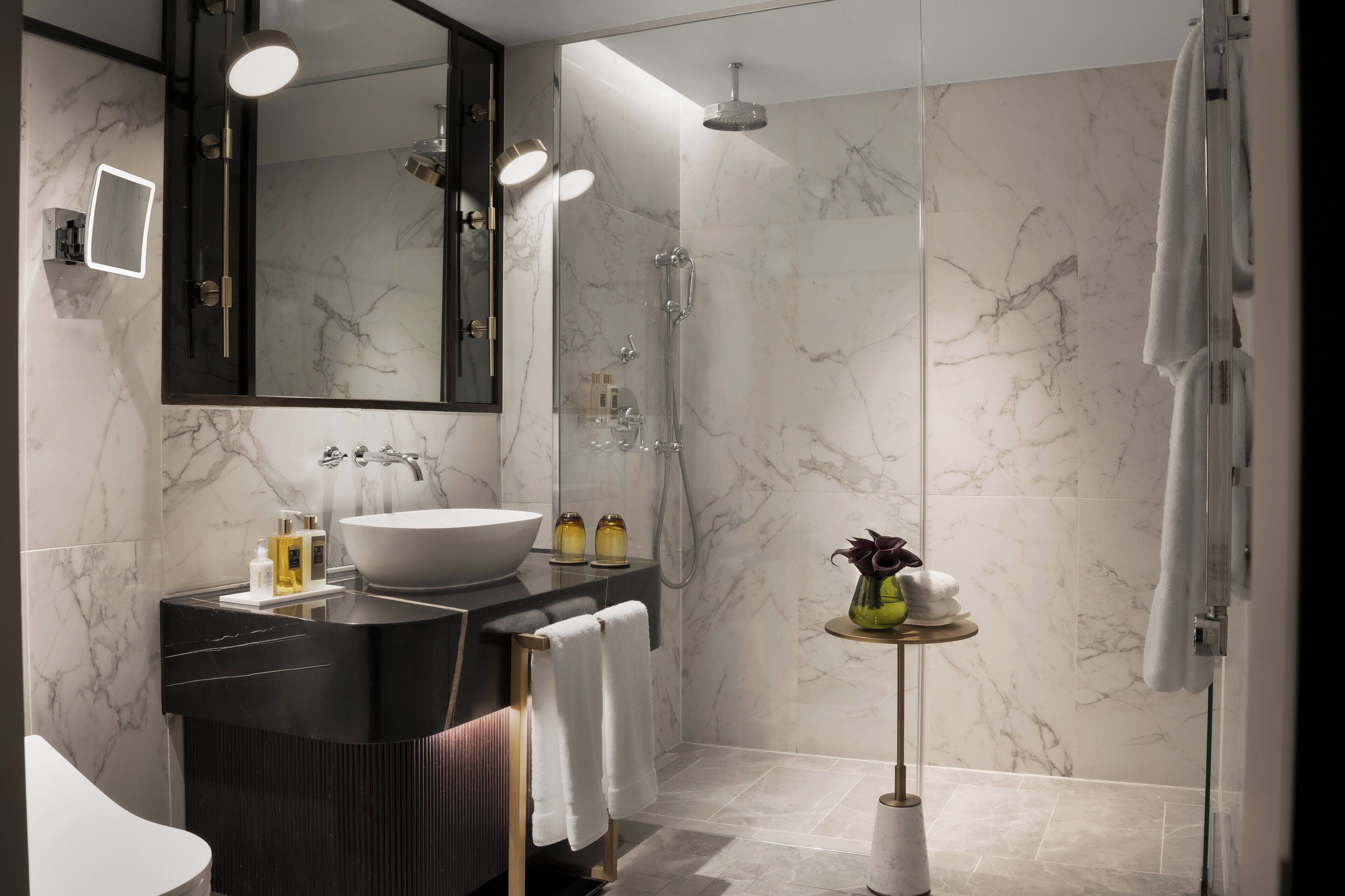 A modern bathroom with a large mirror, a vessel sink, a shower, and a small table with toiletries.