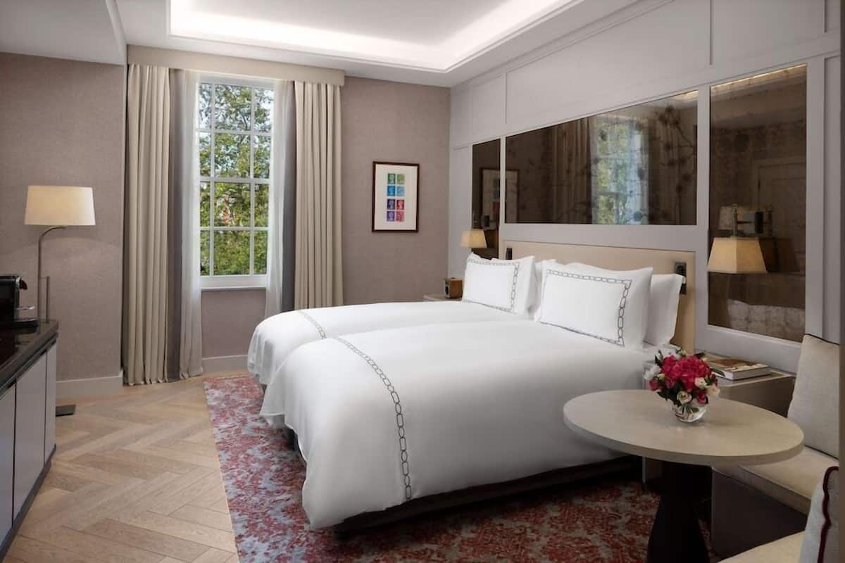 A hotel room with a large bed, a round table with flowers, a desk with a lamp, and a large window with curtains.
