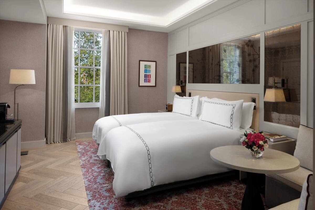 A hotel room with a large bed, a round table with a vase of flowers, a desk with a lamp, and a large window with curtains.