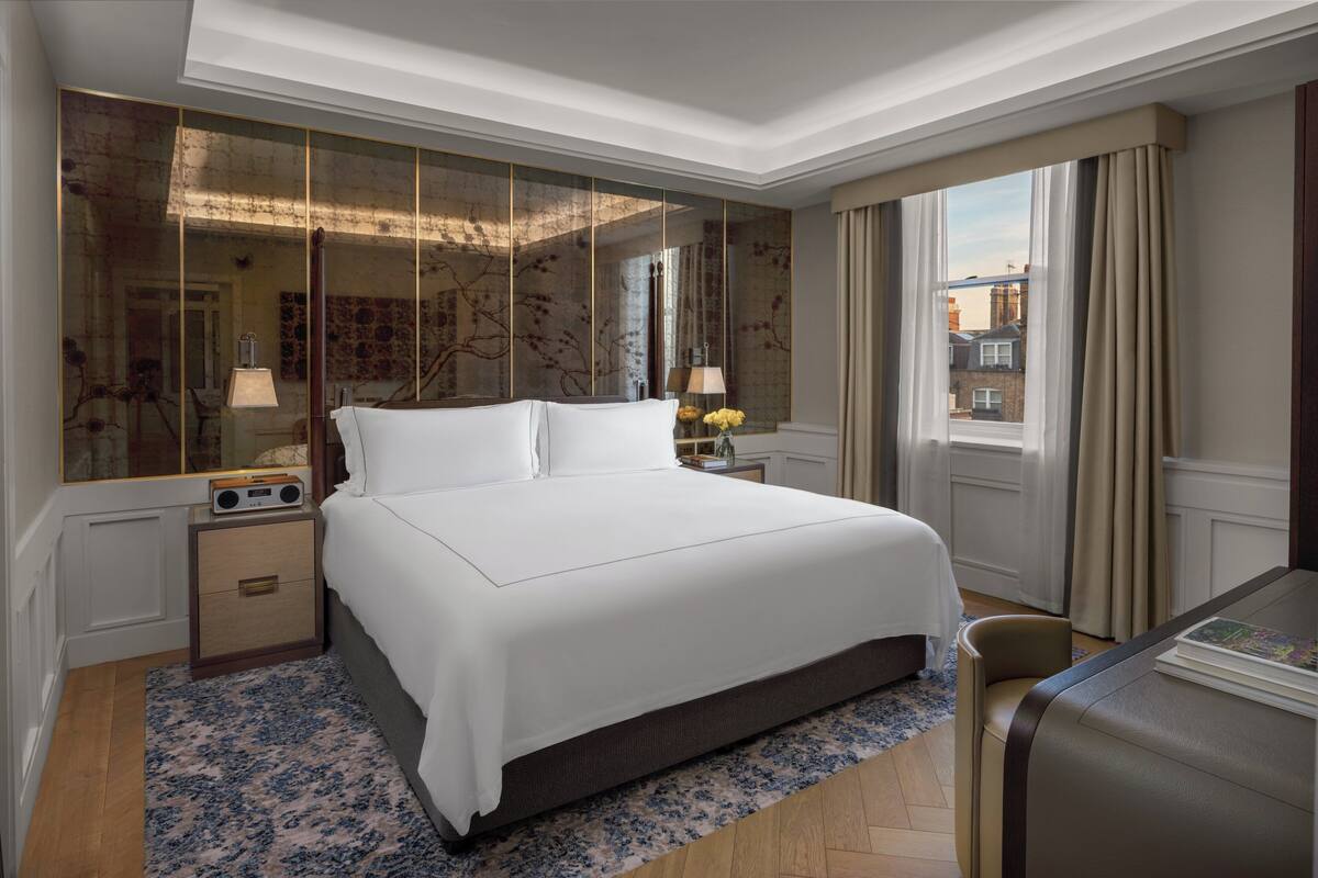 A hotel room with a large bed, a bedside table with a radio, a mirror, and a view of the city through the window.