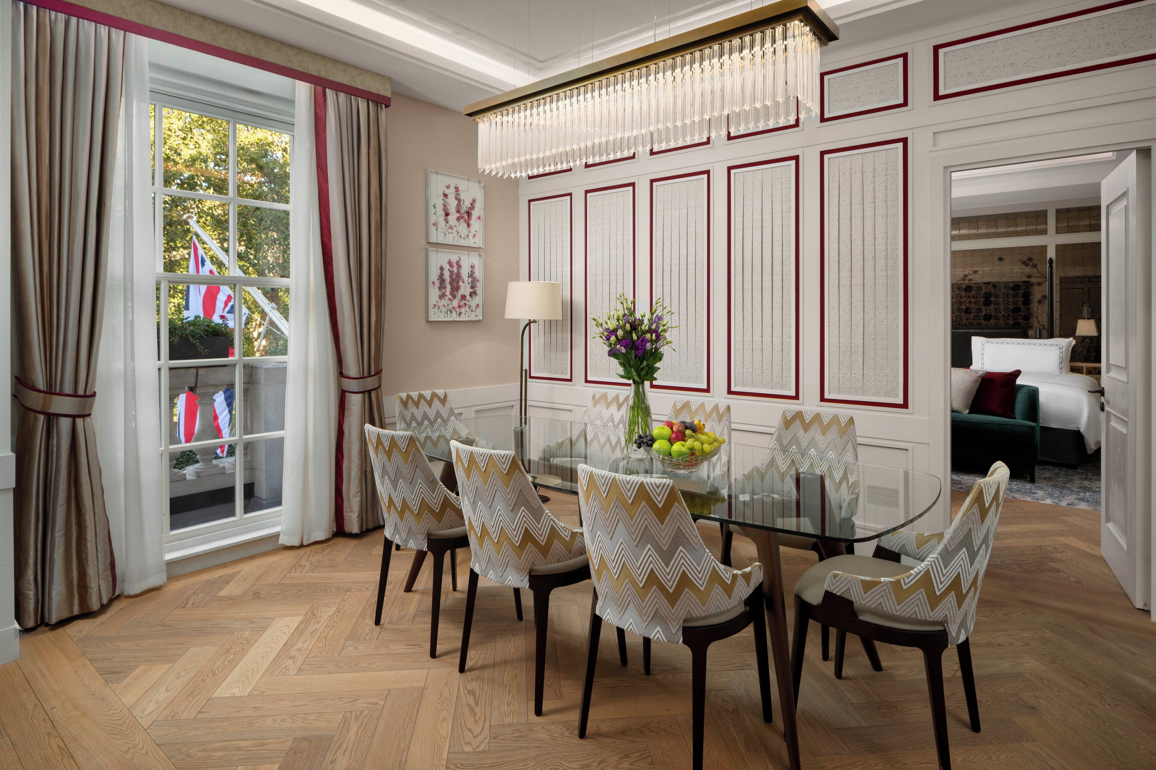 A dining area with a glass table, six chairs with patterned upholstery, a chandelier, and a window with curtains.