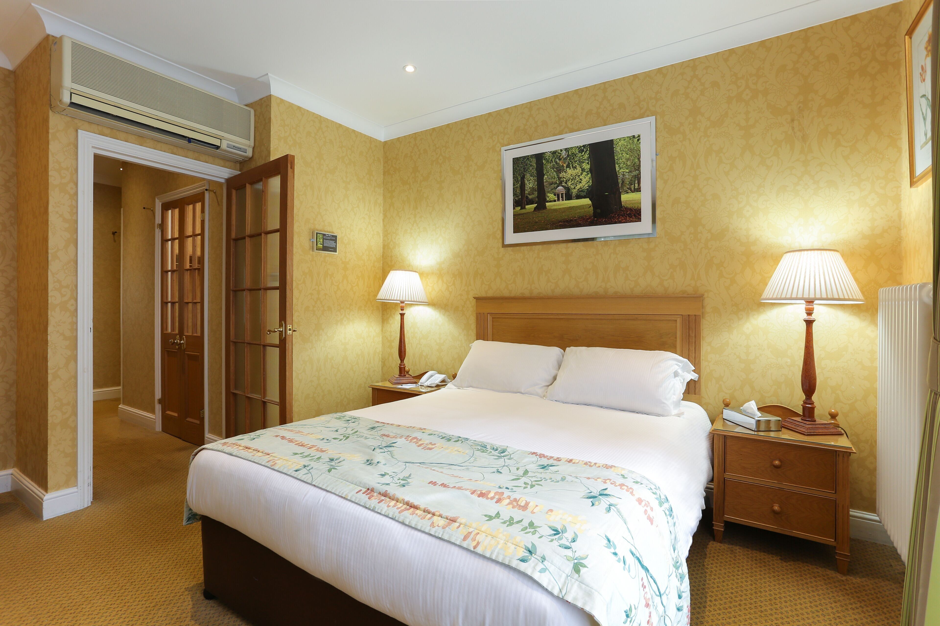 Une chambre d’hôtel avec un grand lit, deux tables de chevet avec des lampes, une tête de lit en bois, un tableau encadré au mur et un appareil de climatisation au-dessus du lit.