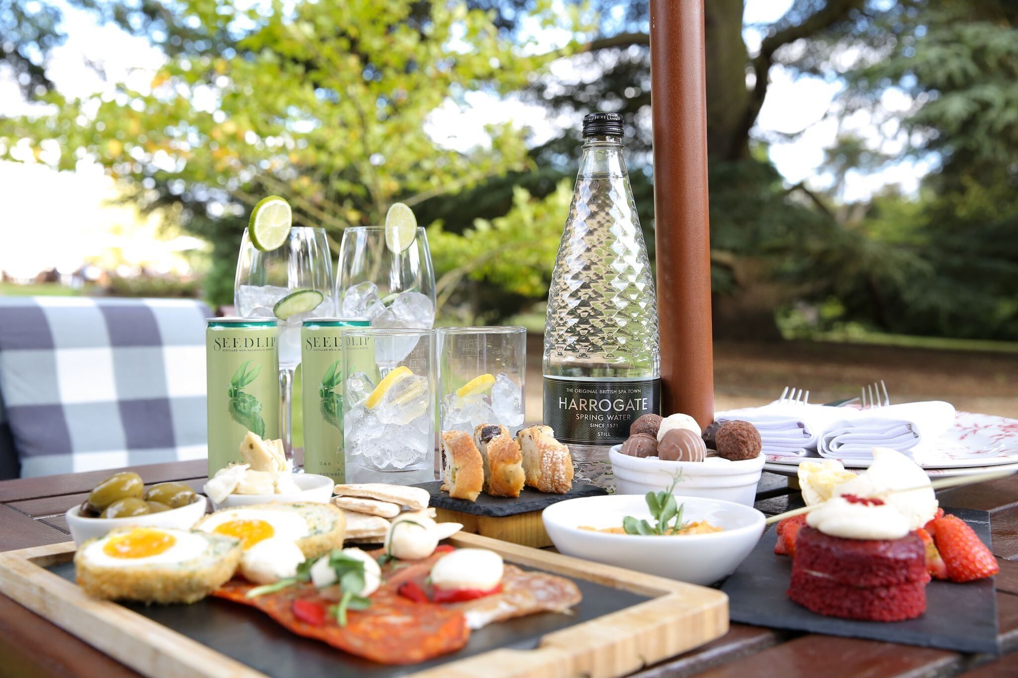 En picknickuppsättning med olika sorters mat och dryck, samt en flaska Harrogate Spring Water, placerad på ett träbord utomhus.