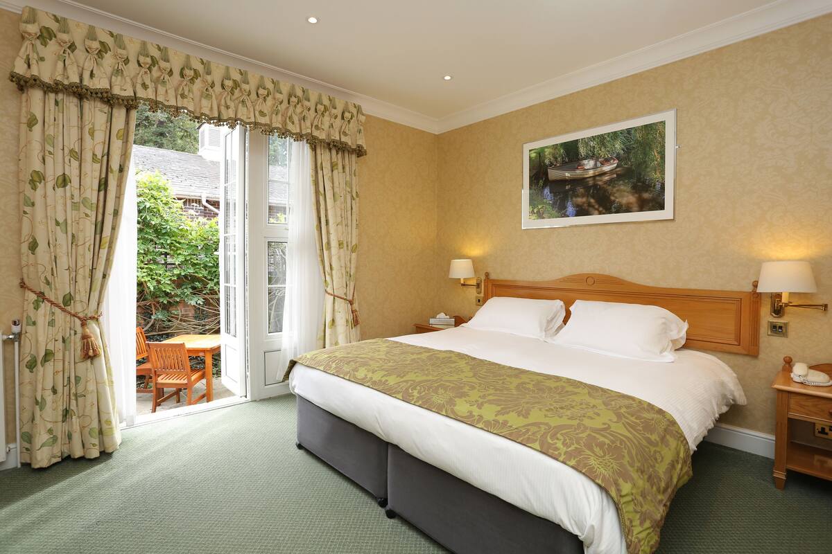 Habitación de hotel con una cama grande, cabecera de madera, dos lámparas de noche, un cuadro en la pared y vistas al exterior a través de una puerta corrediza.