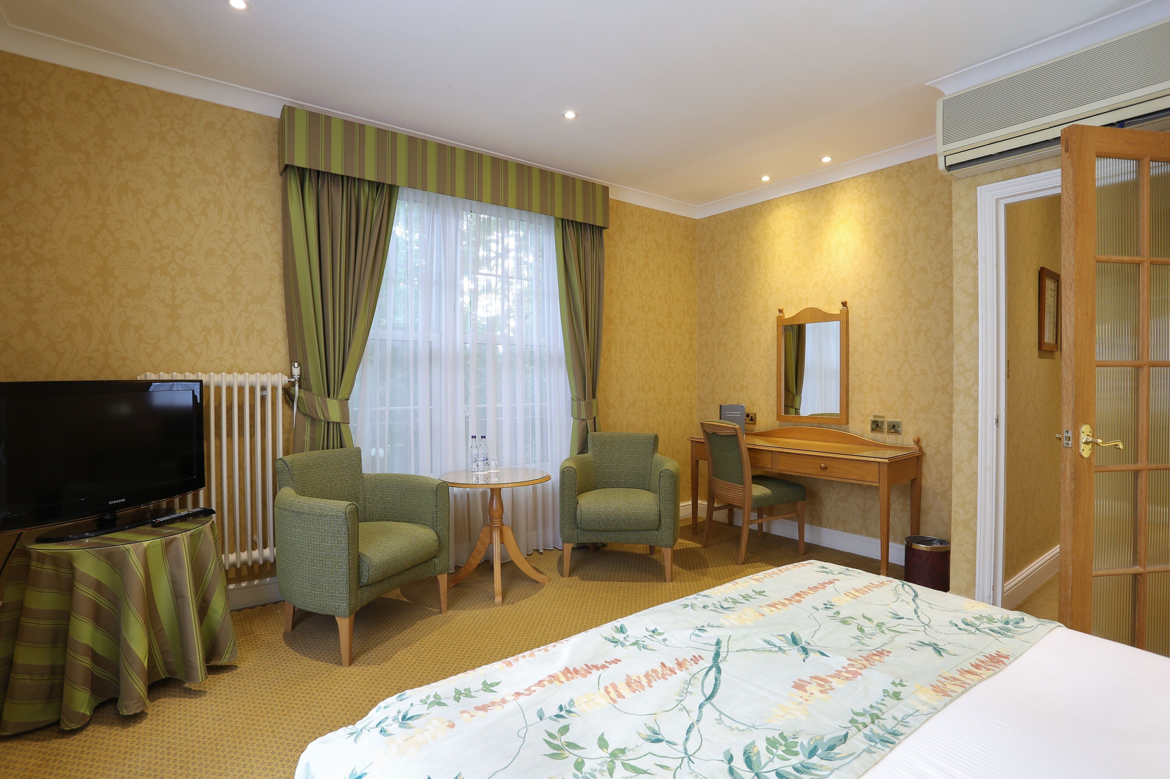 A hotel room with a bed, a television, two armchairs, a desk, and a window with curtains.