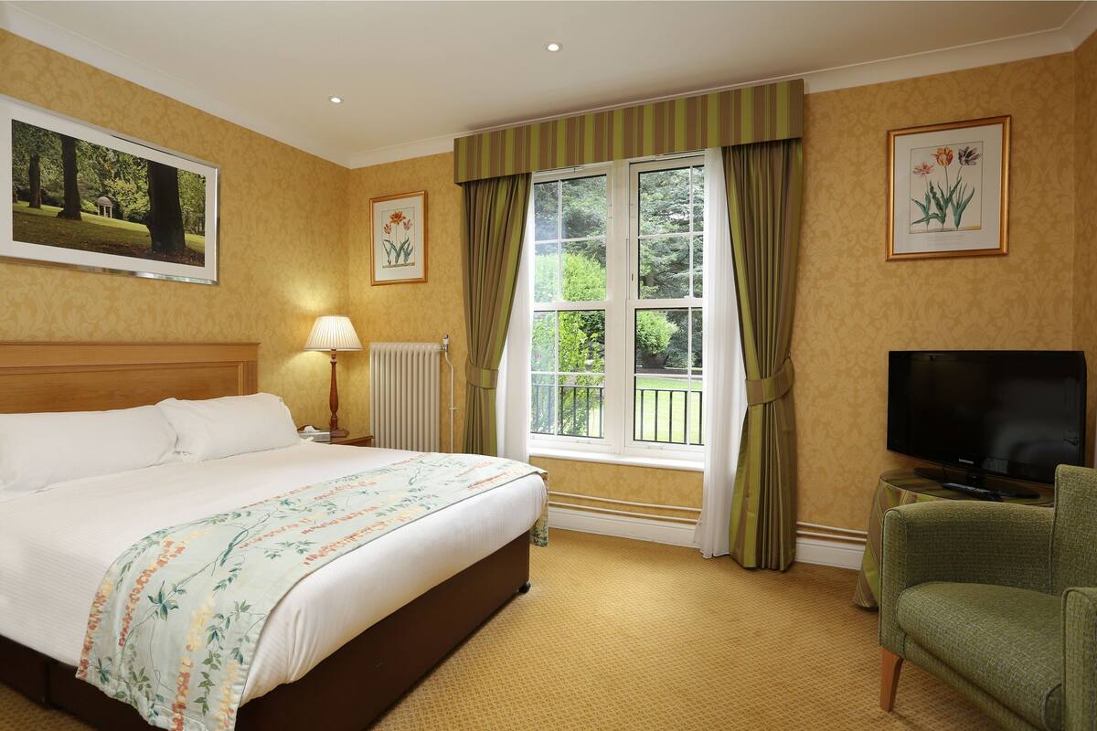 Una habitación de hotel con una cama grande, televisión, un sillón verde y una ventana con cortinas.