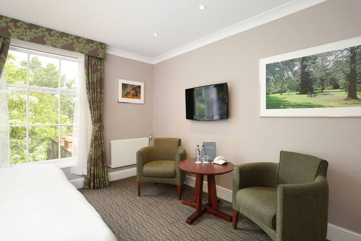 Habitación de hotel con cama, dos sillones, una mesita con teléfono y botellas de agua, un televisor en la pared y una ventana con cortinas.