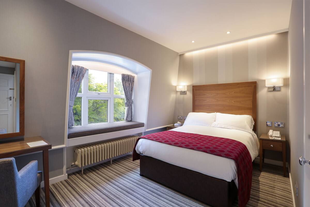 Una habitación de hotel con una cama grande, una ventana con cortinas, una mesita de noche con lámpara y un radiador debajo de la ventana.
