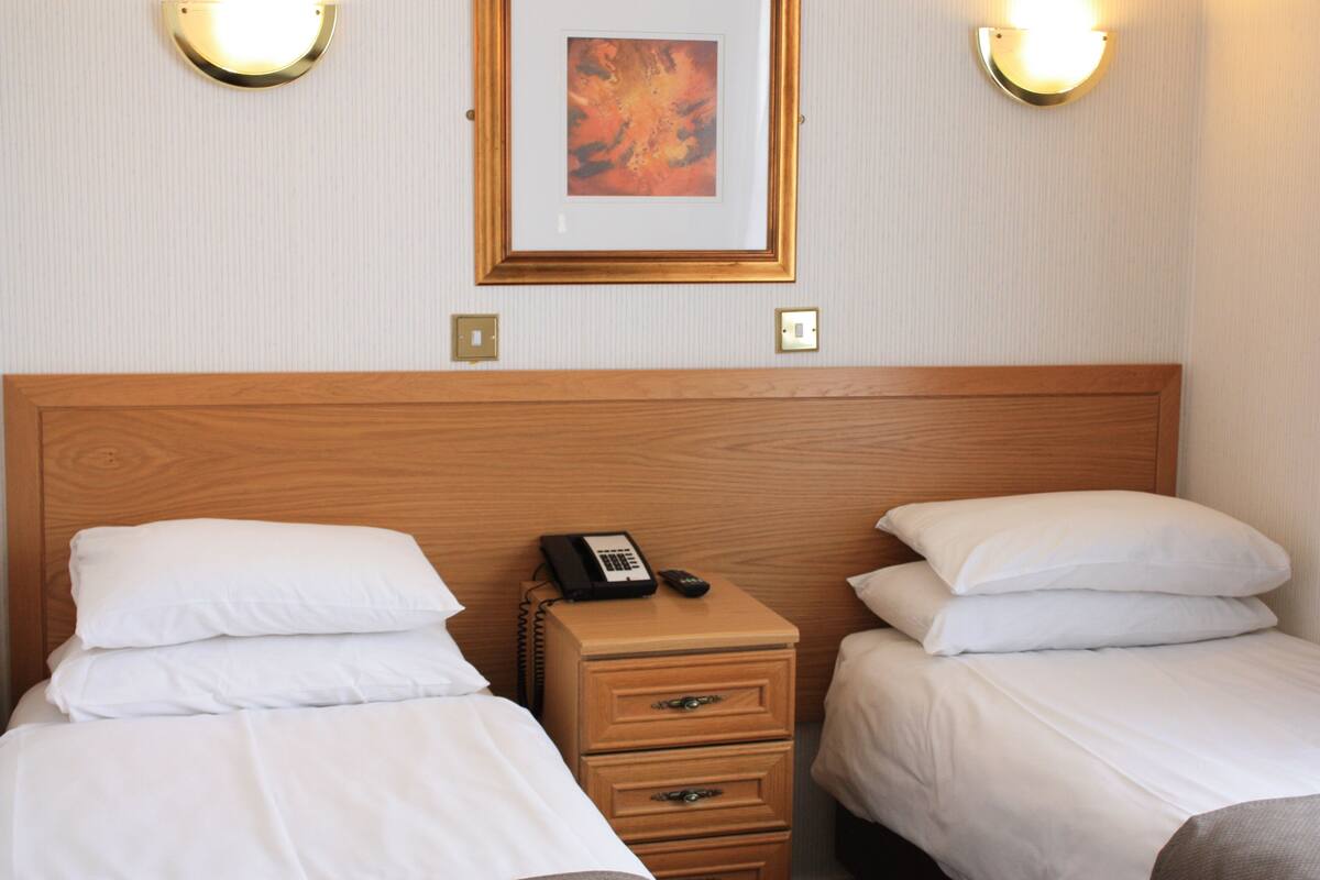 A hotel room with two beds, a wooden headboard, a bedside table with a phone, and wall-mounted lights.