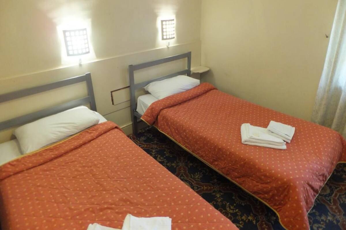 A hotel room with two single beds, each with a white pillow and an orange blanket. There are two bedside tables with lamps, and the room has a carpeted floor.