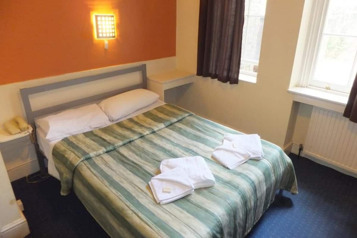 A hotel room with a bed, a nightstand, a lamp, a window with curtains, and a radiator.