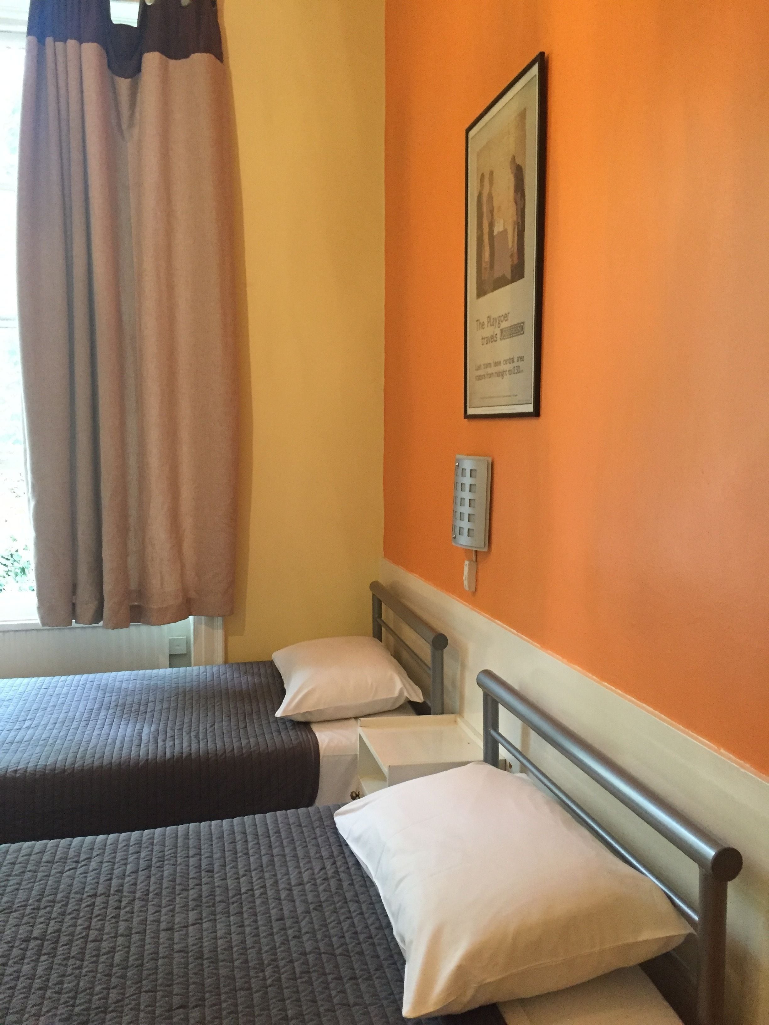 A hotel room with two beds, a window with curtains, an orange wall, and a framed picture.