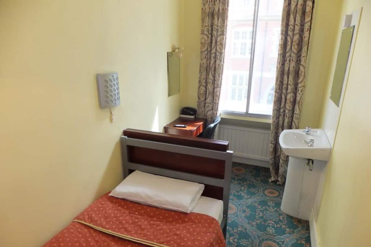 A small, single-bed room with a wooden headboard, a bedside table, a radiator, a window with curtains, and a sink.