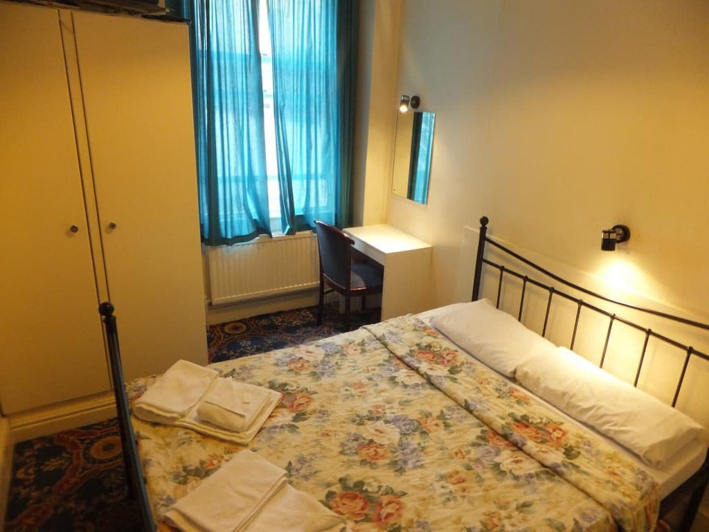A bedroom with a bed, a desk, and a chair. There is a window with curtains, a radiator, and a wardrobe.