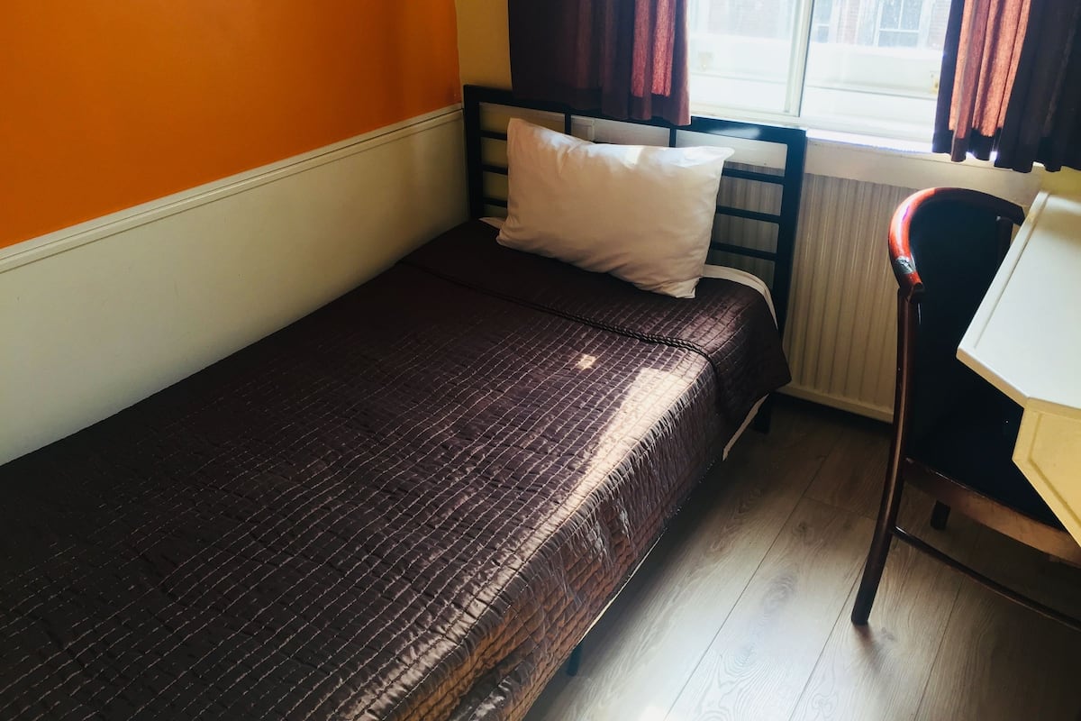 A small, single-bed room with a wooden headboard, a window with curtains, and a radiator.