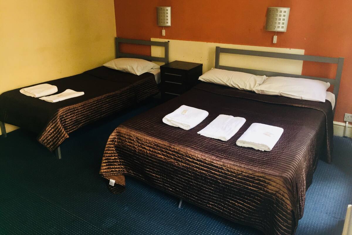 A hotel room with two single beds, each with a brown bedspread and white pillows, and a small bedside table with a lamp.