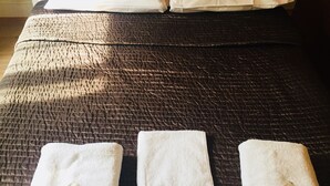 A bed with a dark textured bedspread, two pillows, and three neatly folded white towels with a small tag on each. - Gresham Hotel Bloomsbury (London)