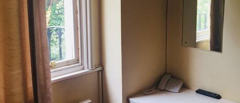 A small room with a window, a white desk, a chair, a television mounted on the wall, and a telephone.
