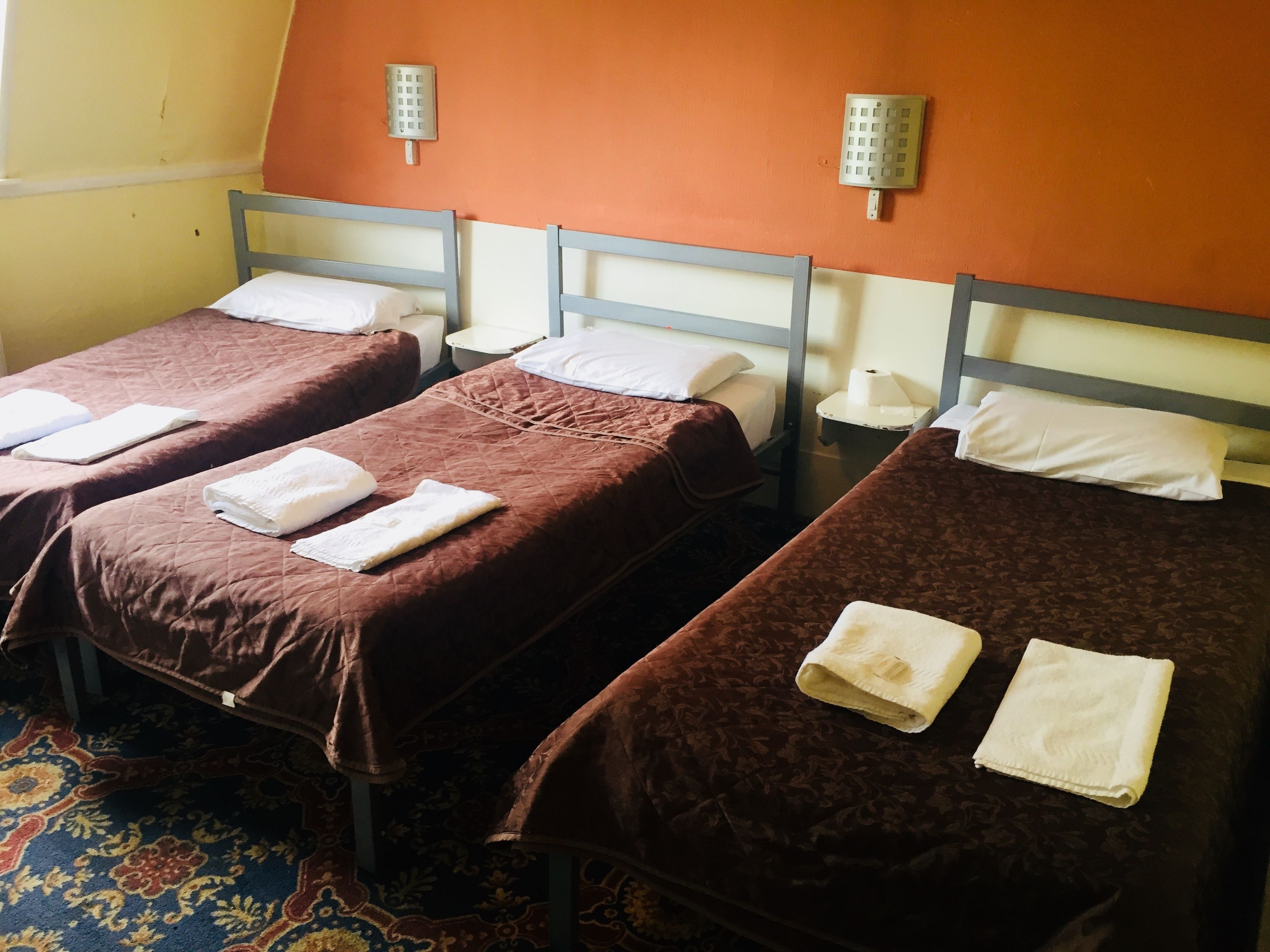 Three single beds with brown linens and white pillows, a patterned carpet, and a wall-mounted light fixture.