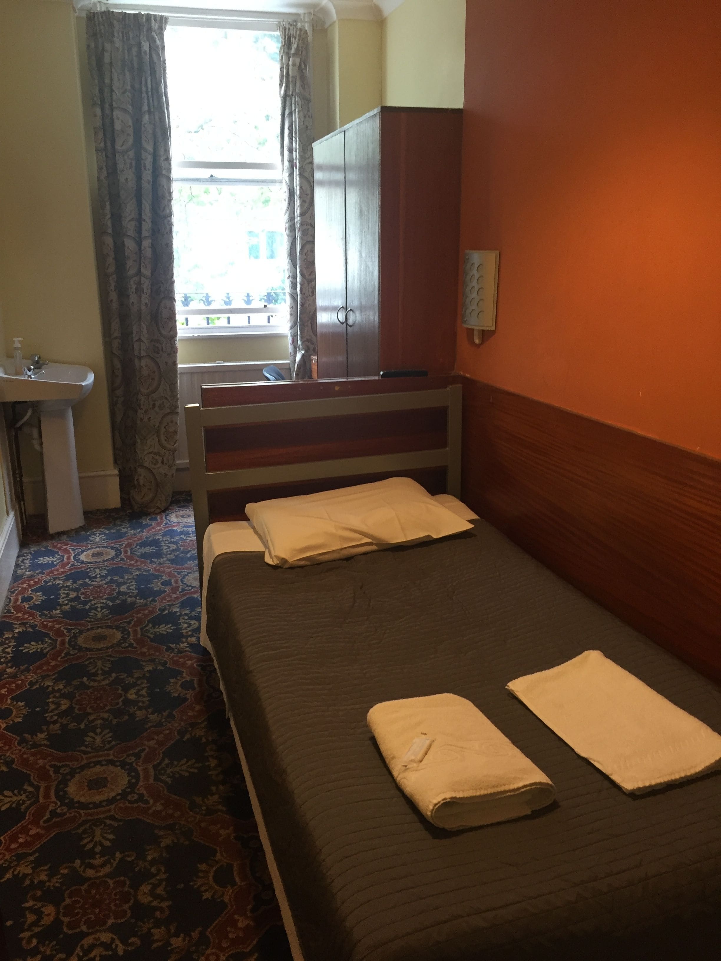 A room with a bed, a window with curtains, a wardrobe, a sink, and a patterned carpet.