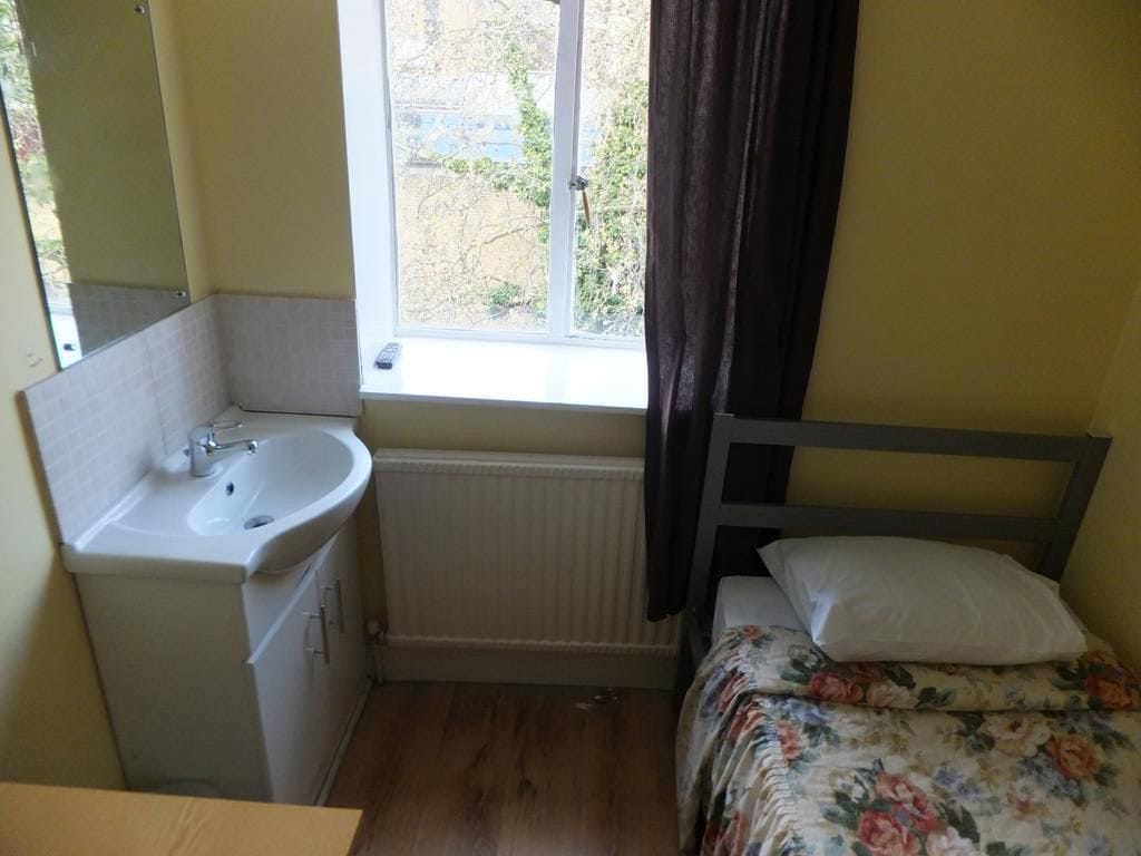 A small, compact room with a single bed, a bedside table, a window with a view of the outside, and a bathroom with a sink and mirror.