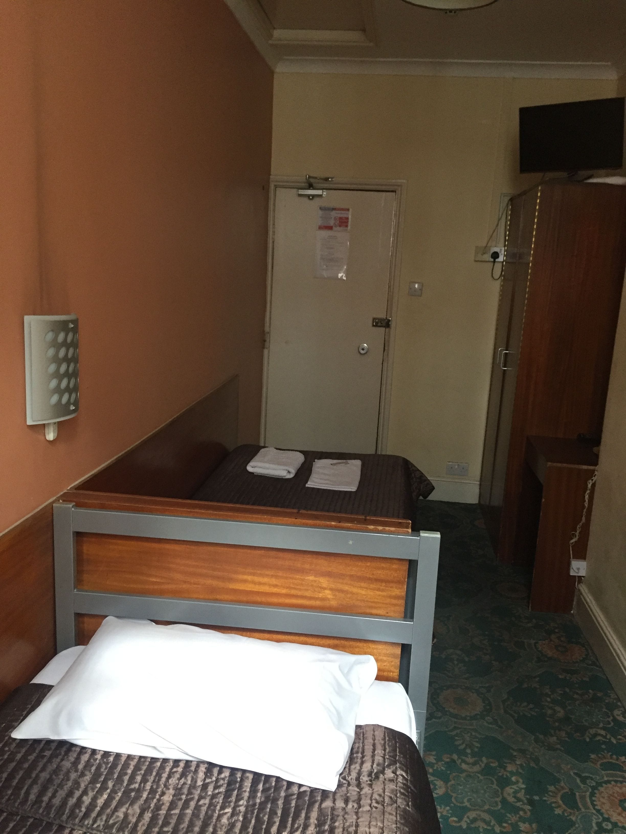 Uma cama de solteiro com cabeceira de madeira, travesseiro branco e toalha branca dobrada. Há uma porta com uma placa, um pequeno armário de madeira e um aplique de parede.