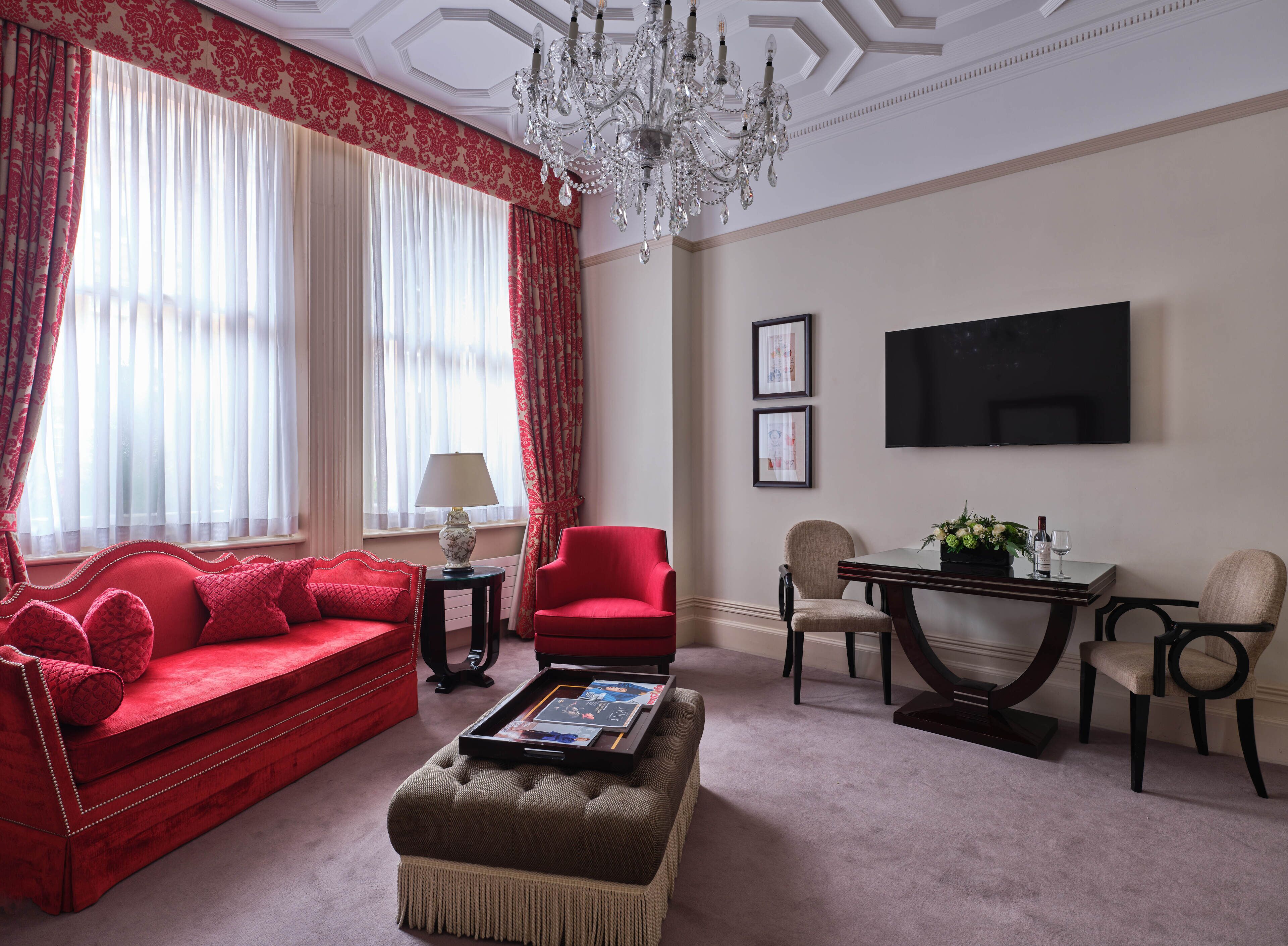 A living room with a red sofa, a red armchair, a coffee table with a tray, a chandelier, a TV on the wall, and a window with sheer curtains.