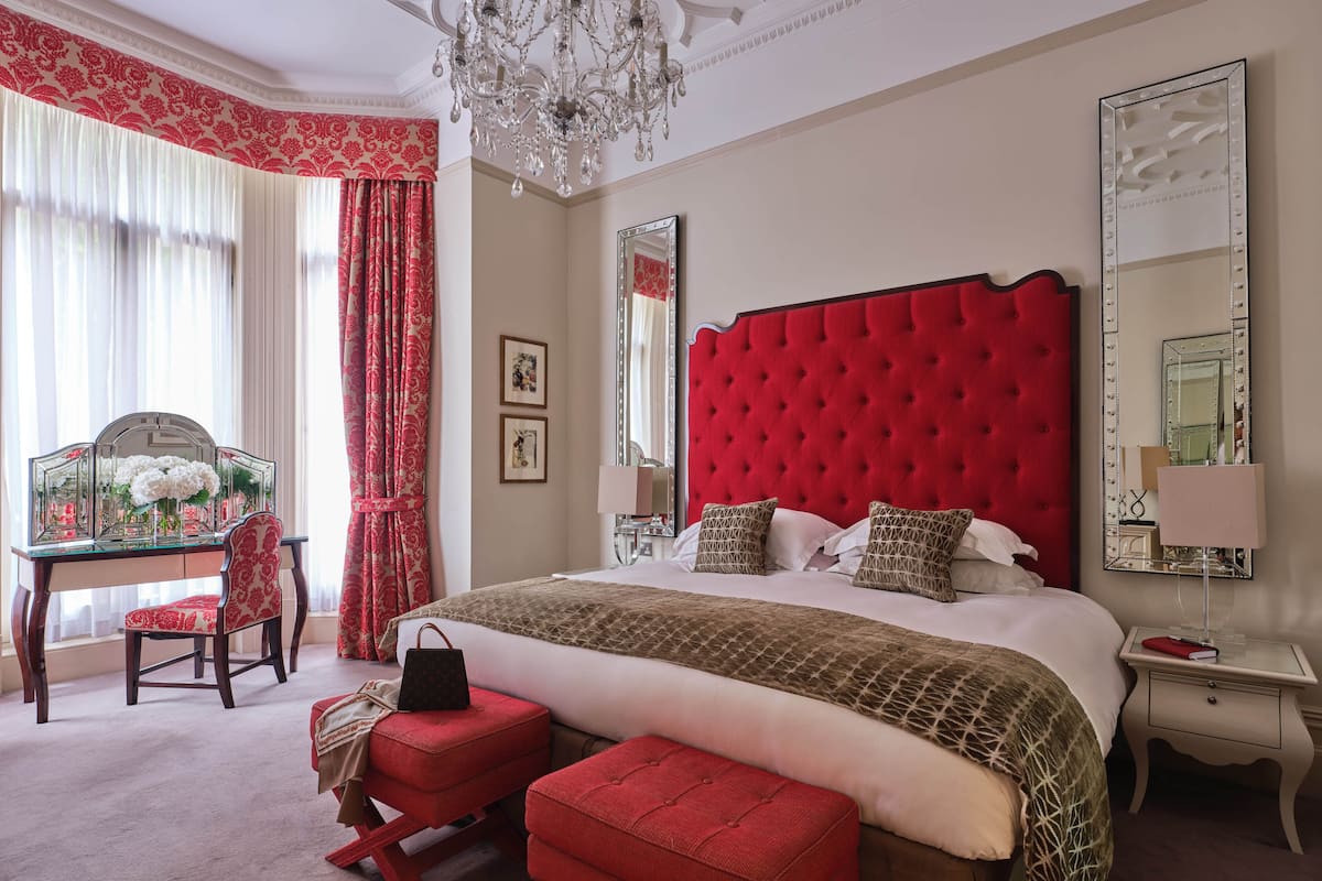 A hotel room with a large bed, a vanity with a mirror, a red upholstered chair, and a red ottoman.