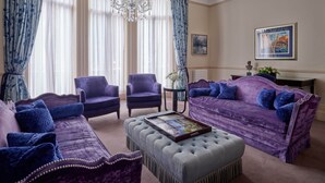 A room with a purple sofa, two armchairs, a chandelier, and a framed picture on the wall.
