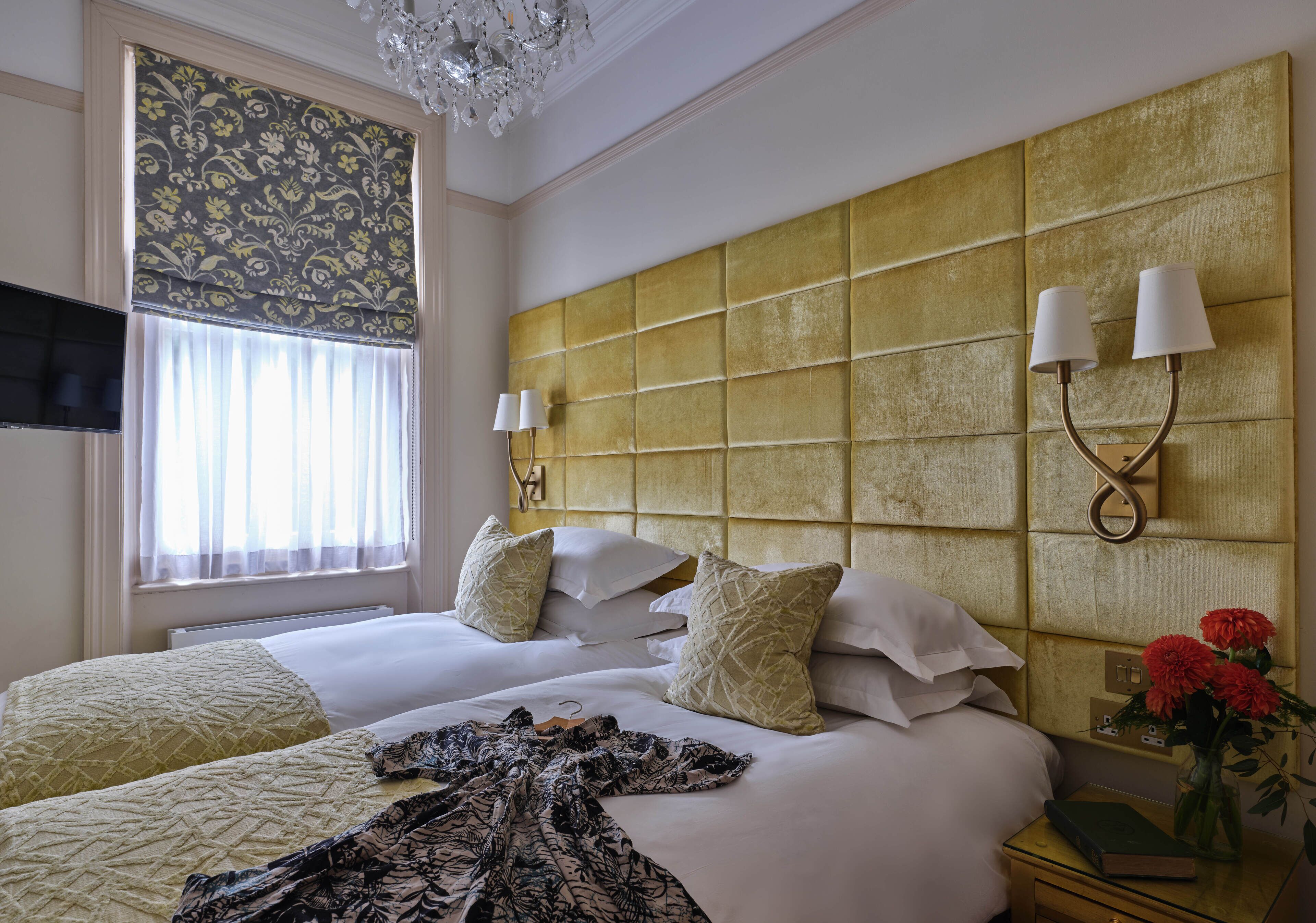 Habitación de hotel con dos camas, un cabecero grande, apliques de pared, un televisor de pantalla plana y una mesita de noche con un libro y flores.
